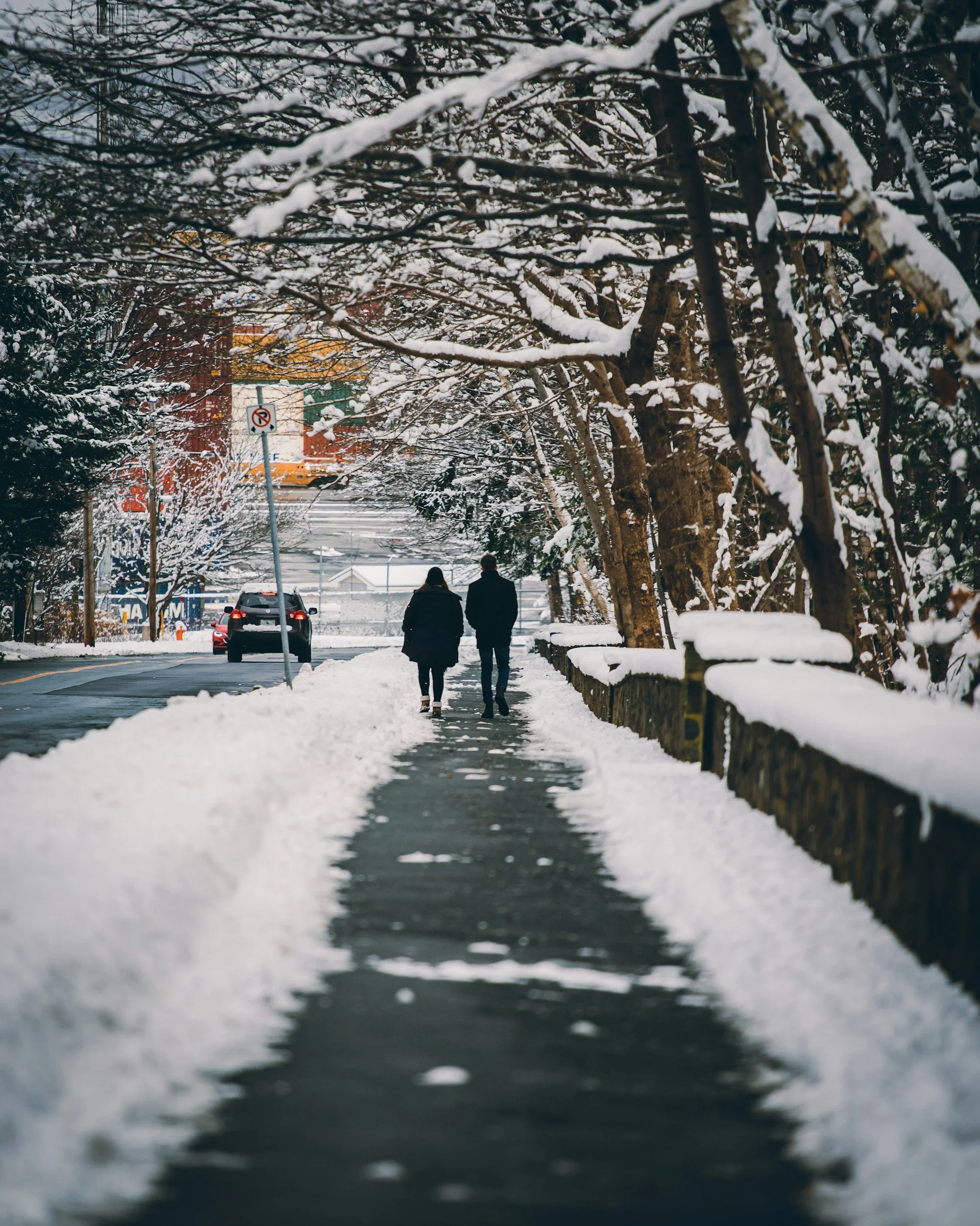 Zwei Menschen gehen im Schnee auf einem Gehweg, über denen sich schneebedeckte Bäume erstrecken.
