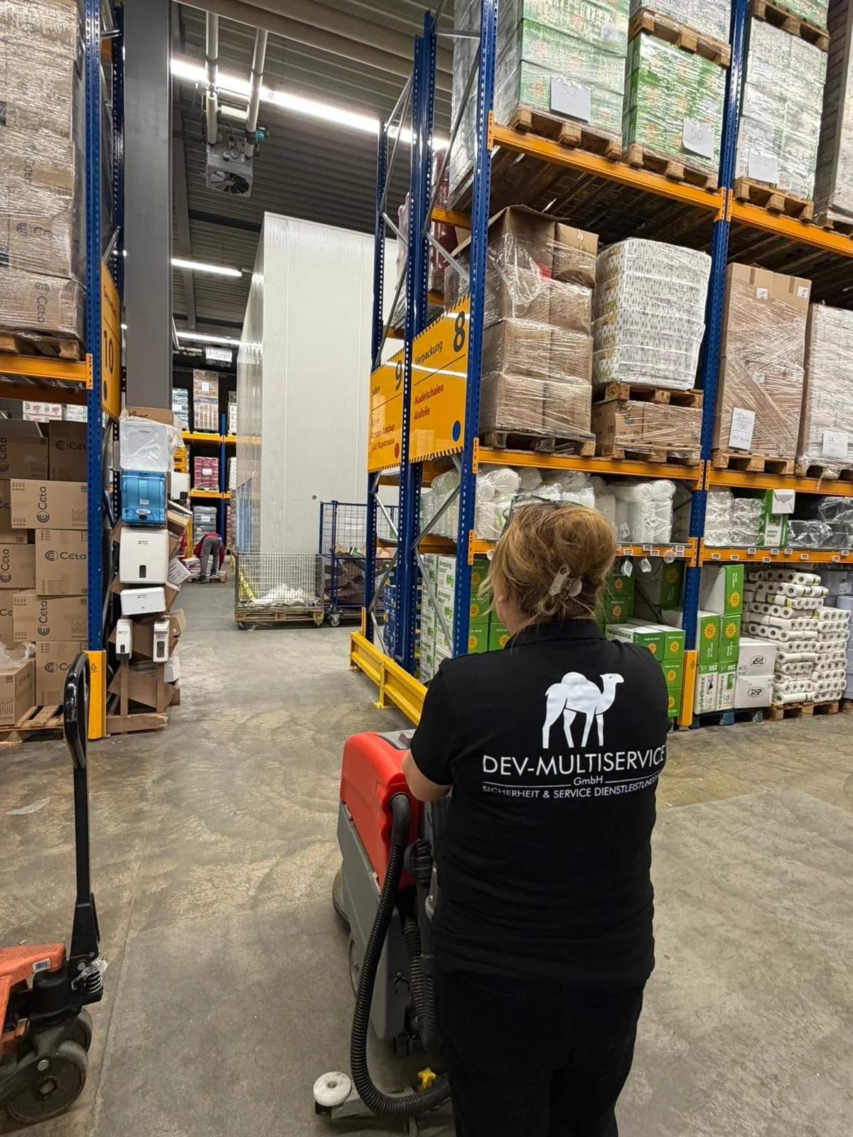 Mitarbeiterin mit DEV-MULTISERVICE-Shirt beim Reinigen eines Lagerhallenbodens in einem großen Lager mit hohen Regalen voll mit Verpackungsmaterialien und Waren.