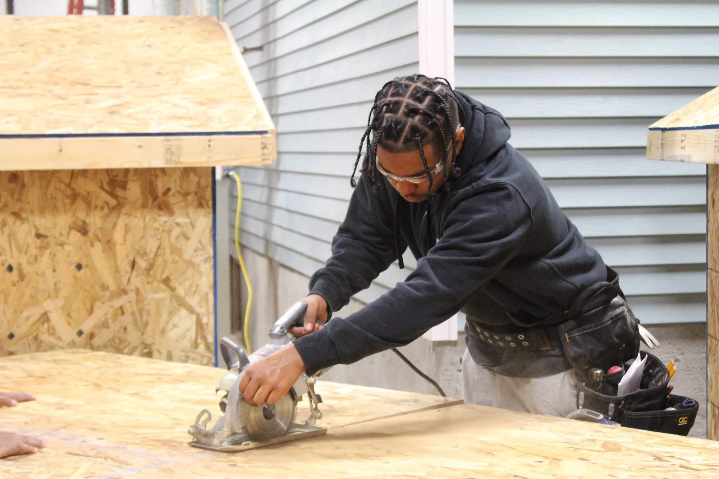Person using a circular saw to cut a piece of plywood at a construction site.
