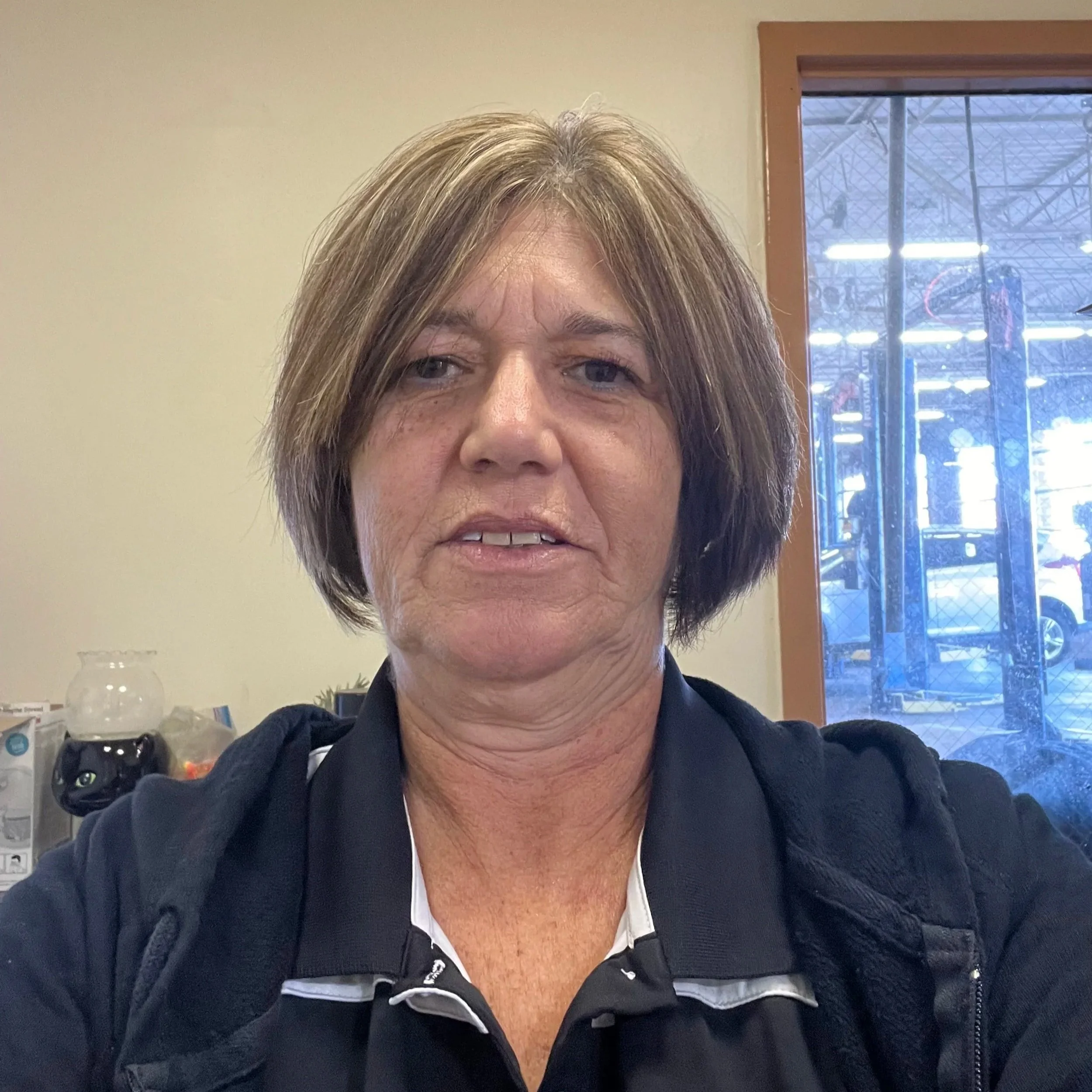 A woman taking a selfie indoors, with a window showing a view of a parking lot and gym equipment outside.