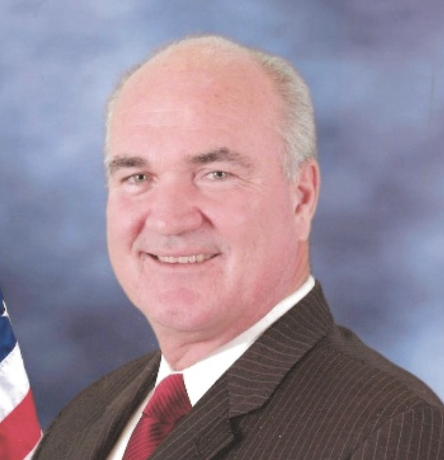 A middle-aged man with short gray hair, dressed in a dark pinstripe suit, white shirt, and red tie, smiling at the camera against a background with an American flag partially visible.