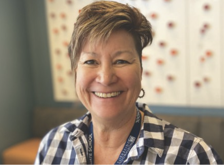 A smiling woman with short hair wearing a checkered shirt and a lanyard around her neck, standing indoors with a colorful wall in the background.