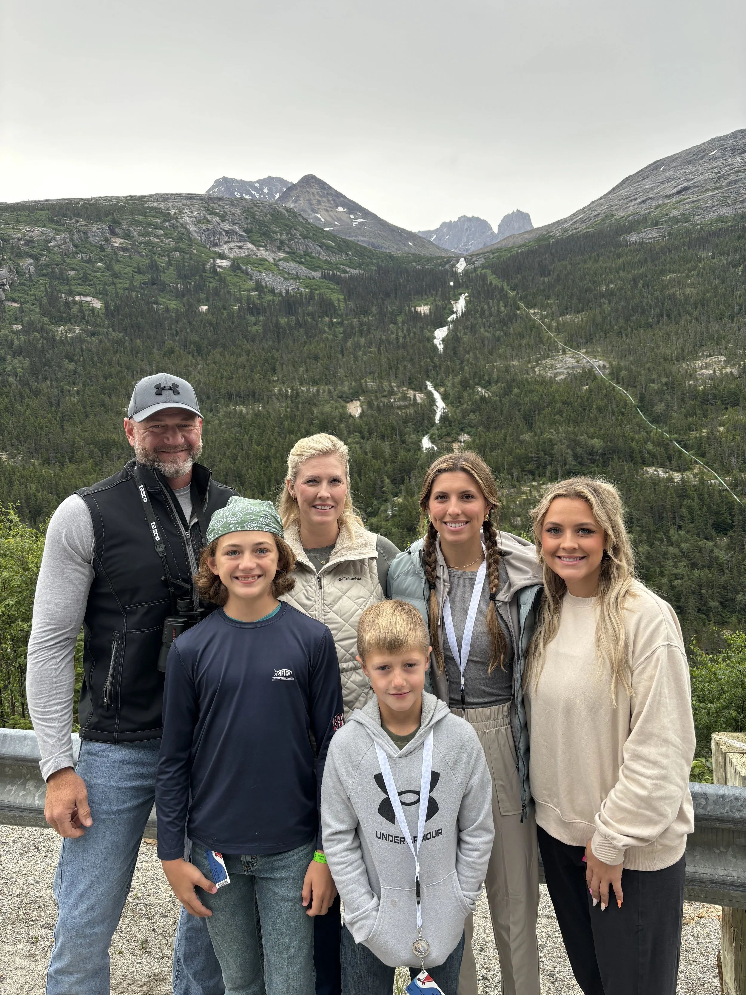 A group of seven people, including two children and five adults, standing outdoors in front of a mountainous landscape with lush green trees and waterfalls, during daytime.