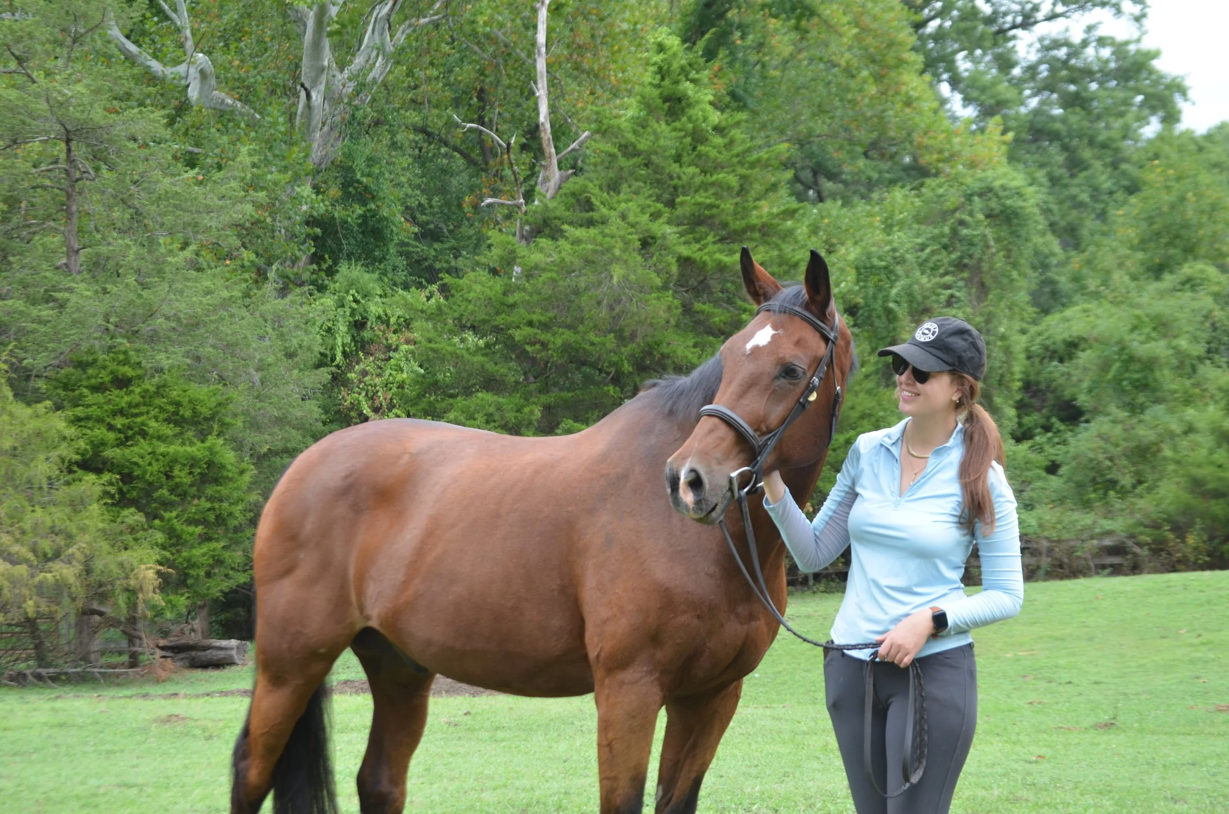 A woman with long hair, wearing a black cap, sunglasses, a light blue jacket, and dark pants, is standing on a grassy field holding a brown horse with a black mane and tail, surrounded by green trees.