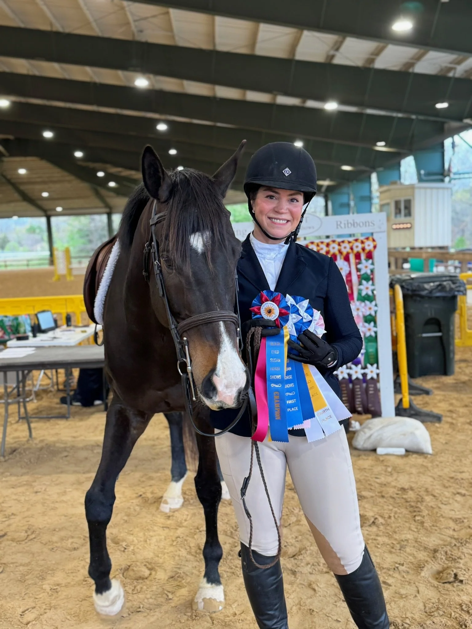 A smiling young woman in riding attire holding a horse's bridle, displaying award ribbons in an indoor equestrian arena.