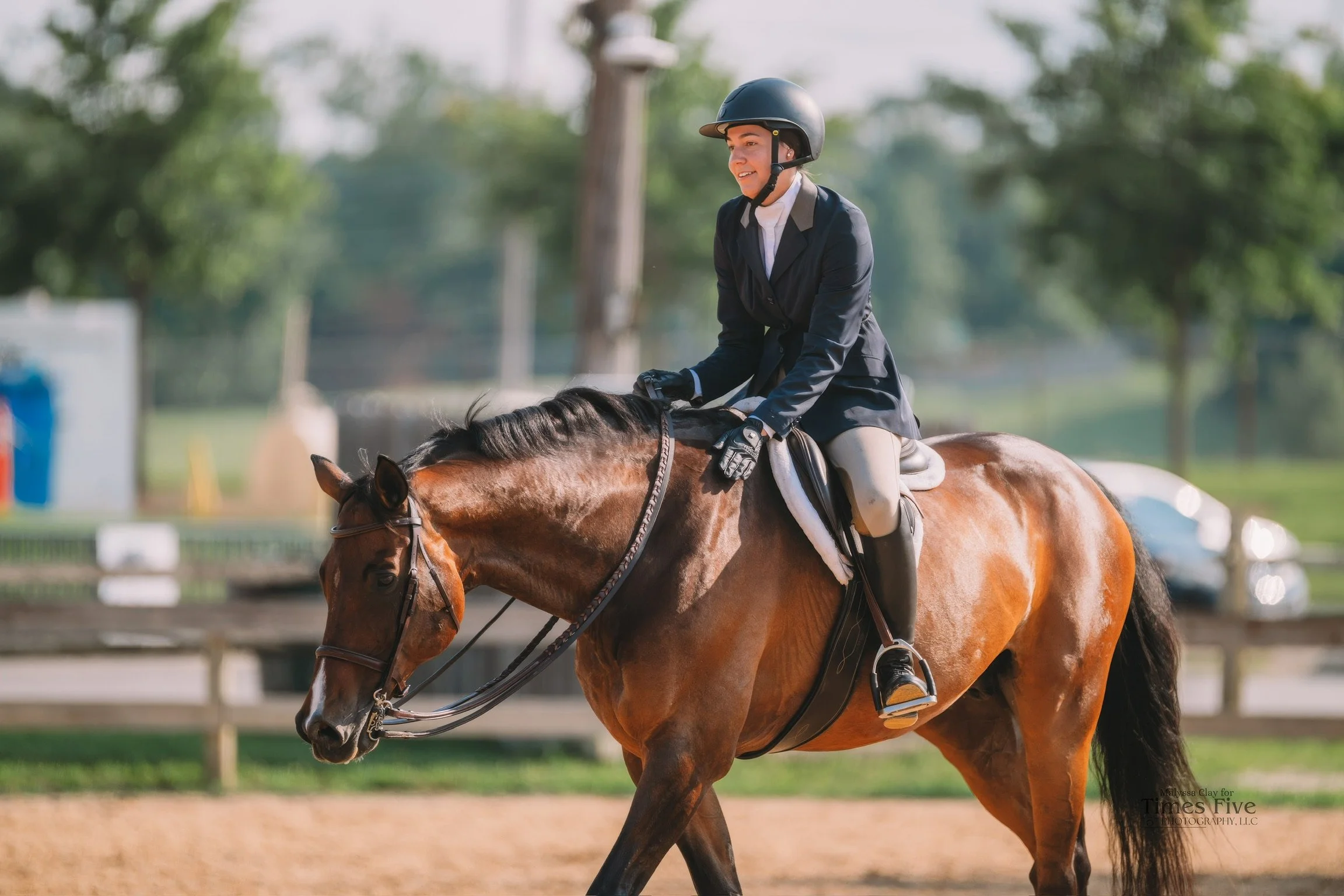 A woman in formal riding attire, including a helmet, is riding a brown horse outdoors on a sunny day.