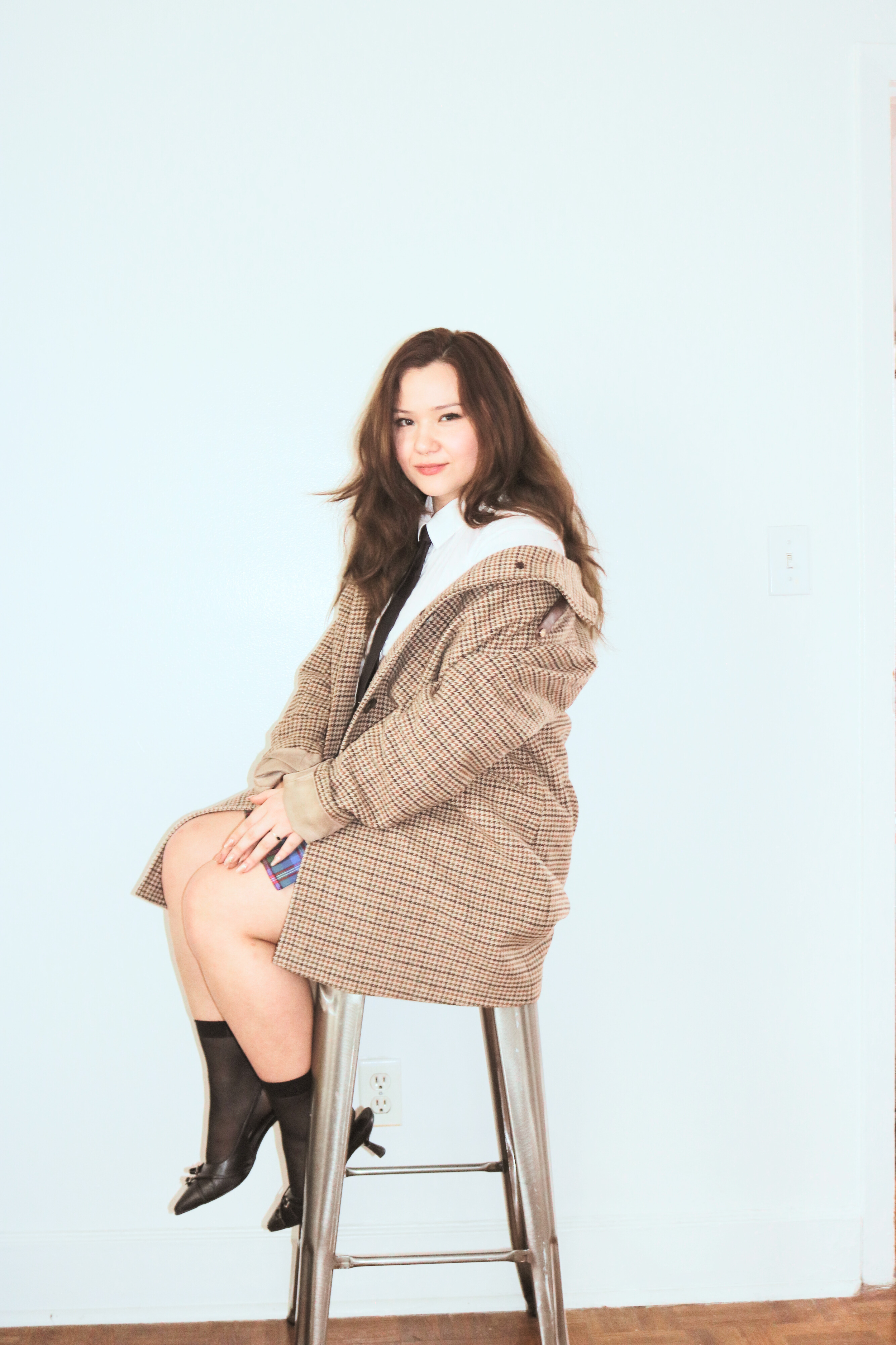 A young woman with long brown hair, sitting on a high stool, wearing a plaid coat, white shirt, black tie, patterned skirt, black socks, and black shoes, against a plain light background.