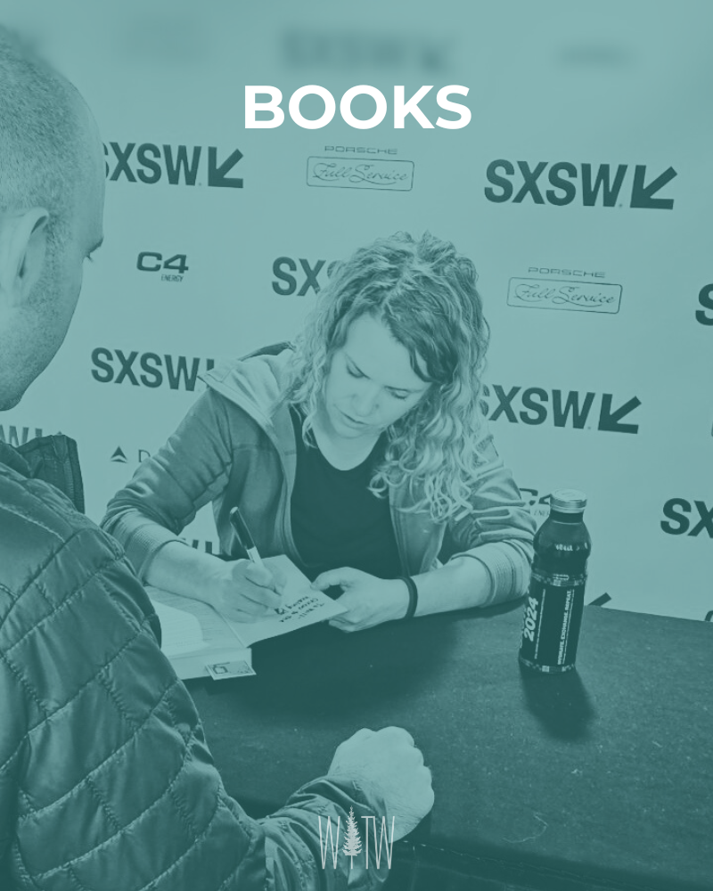 A woman is sitting at a table signing a book while a man is seated across from her with his hands on the table. There is a bottle on the table. The background has logos for SXSW, Porsche, and Fuel Service, indicating this is an event or signing session.