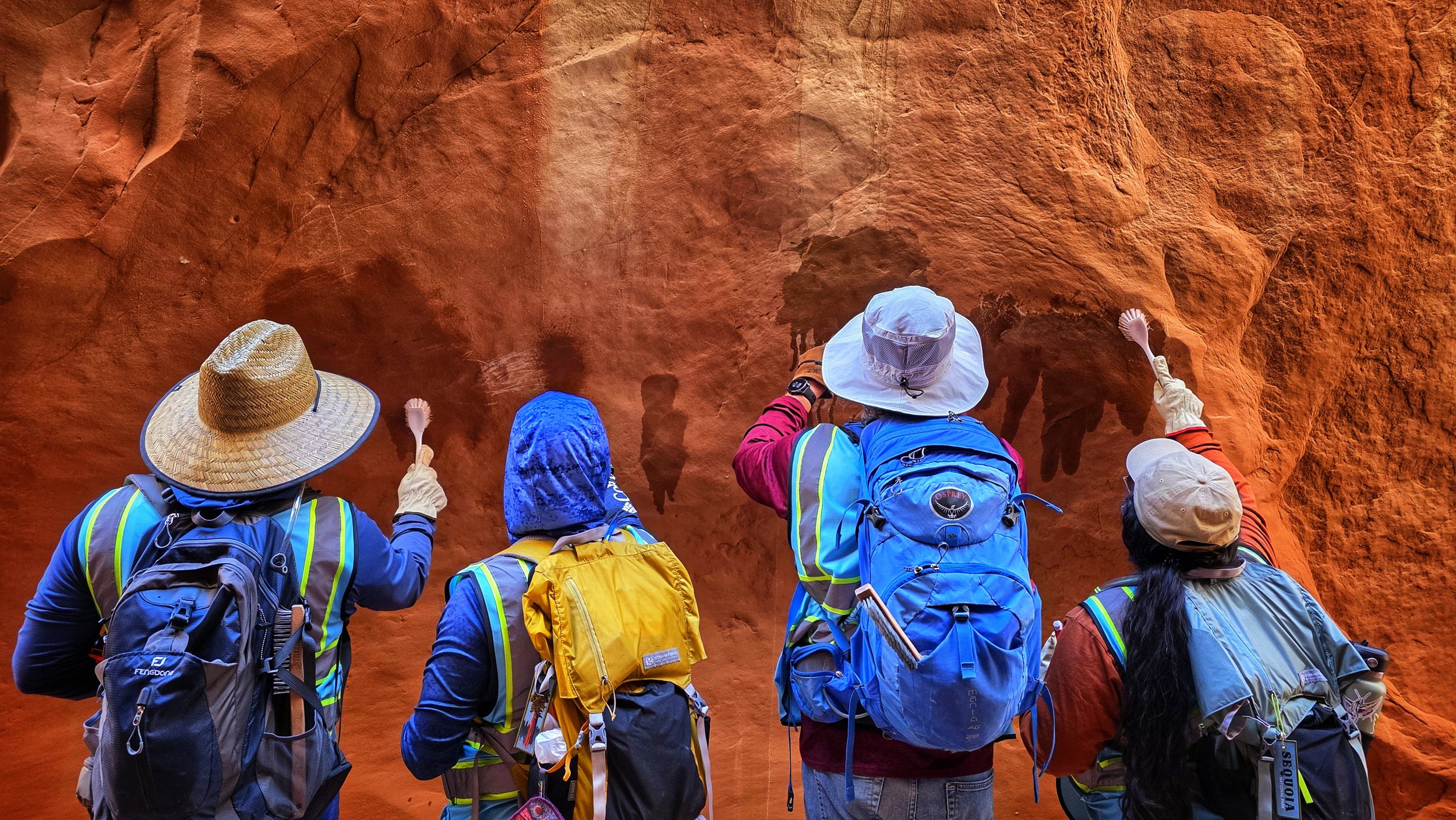 Four hikers with backpacks and hats exploring a red rock wall, with some pointing or touching the surface.