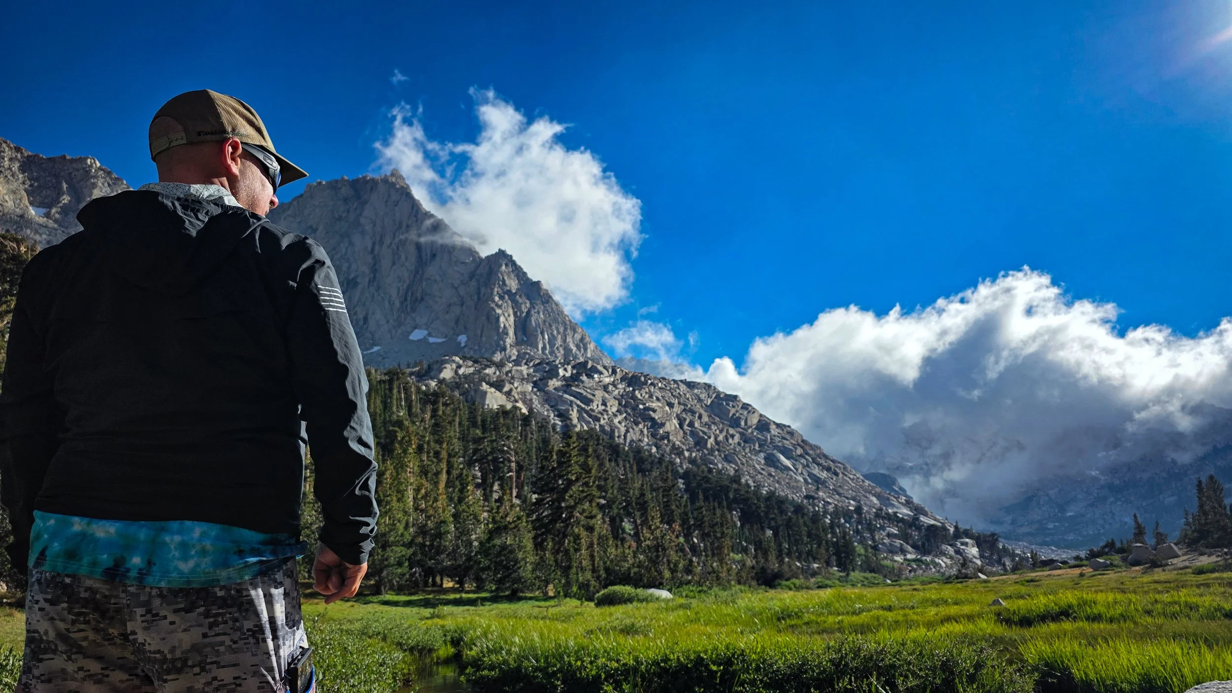 A man dressed in a black jacket, camouflage shorts, and a baseball cap stands outdoors in a lush green field, looking toward towering rocky mountains with patches of snow, dense trees, and partly cloudy sky in the background.