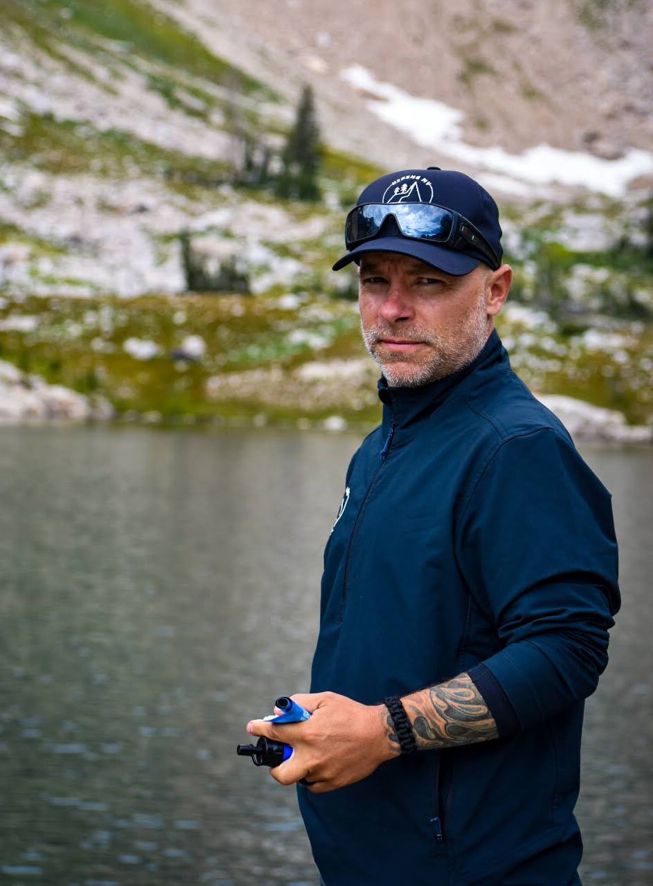 A man stands outdoors near a lake, wearing a dark jacket, sunglasses on his head, and a baseball cap. He has a serious expression and tattoos on his left arm, with a natural landscape of trees and a rocky hillside in the background.