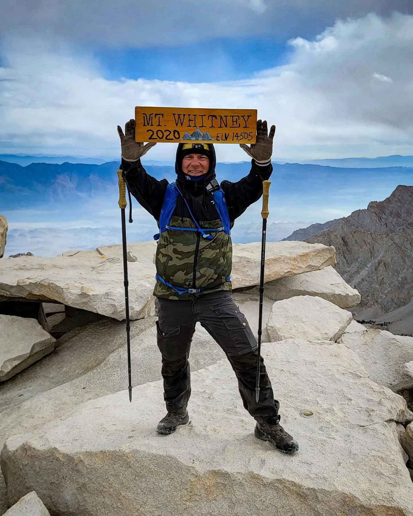 Hiker standing on rocky mountain summit holding a sign that reads 'Mt. Whitney 2020 Elev 14,505' with rugged mountain landscape in the background.