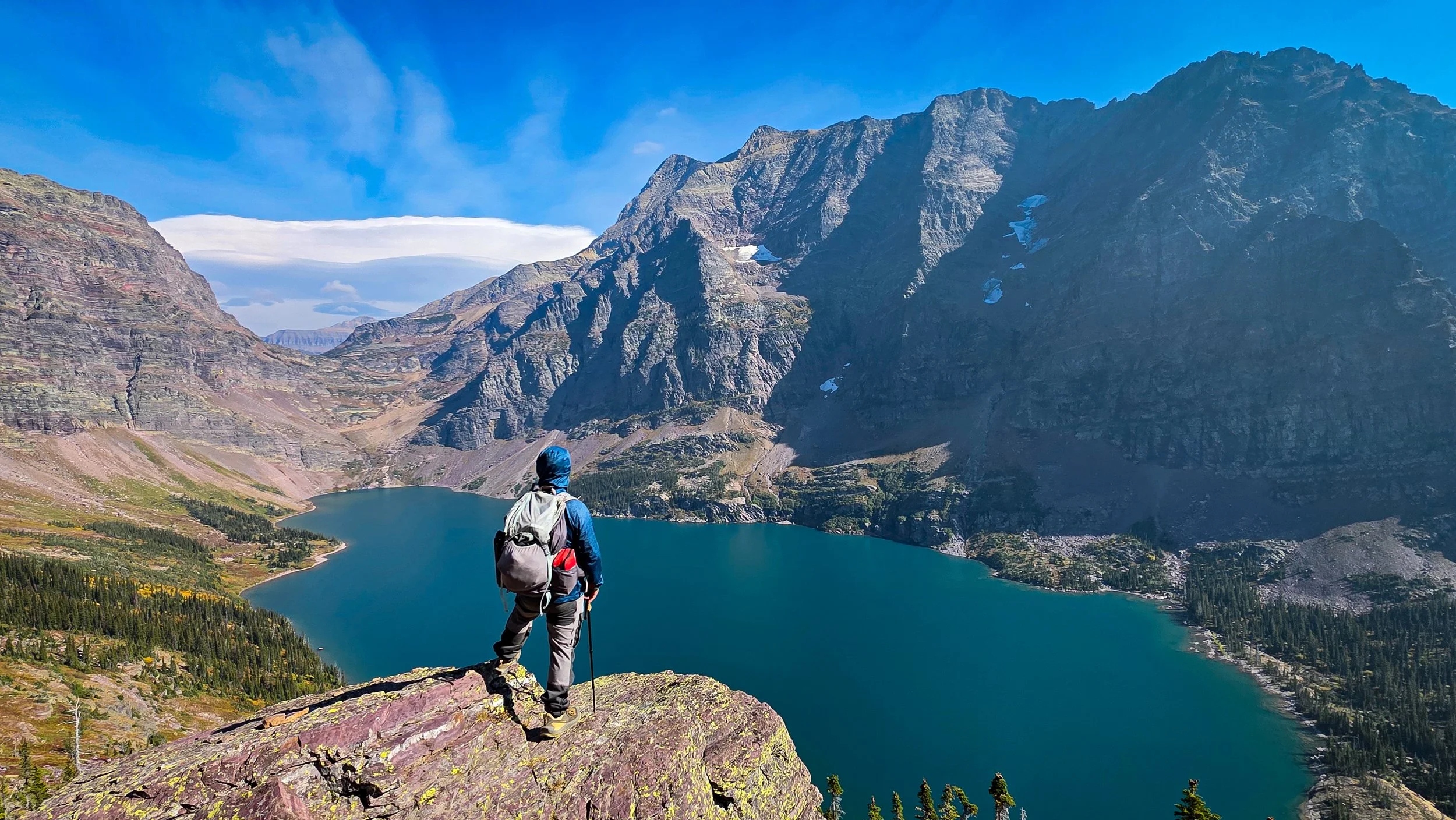 A hiker stands on a rocky ledge overlooking a deep blue mountain lake, surrounded by steep, rugged mountains under a clear blue sky.