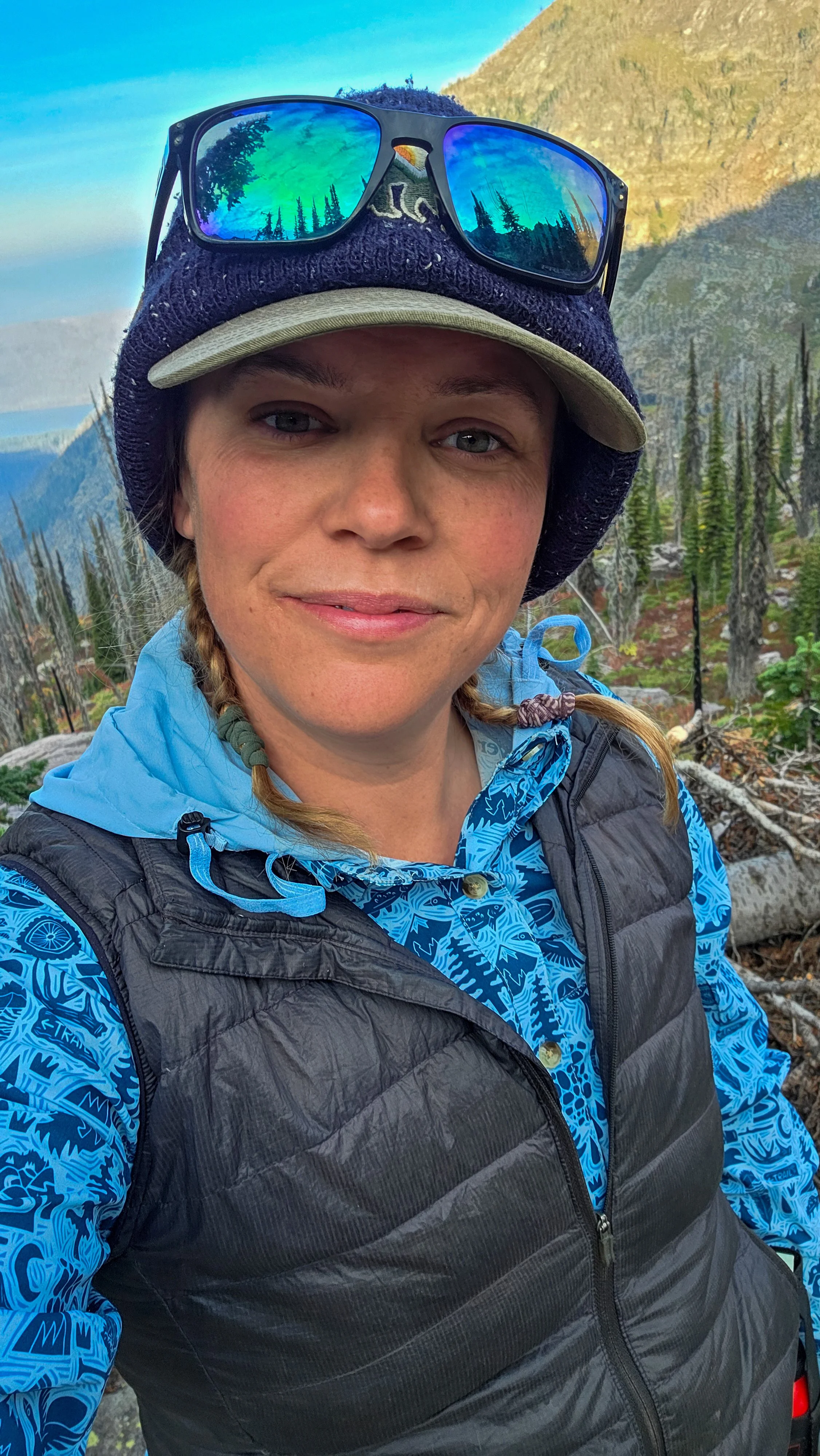 A woman outdoors in a mountainous forest area, wearing a blue patterned jacket, a black puffy vest, a beige hat, and sunglasses with reflective lenses showing trees and sky.
