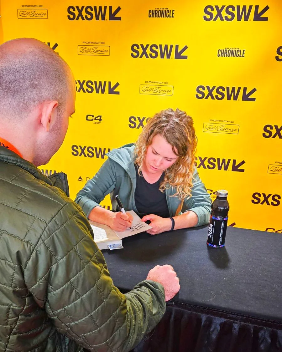 A woman with curly blonde hair signing an autograph at an event with a yellow backdrop featuring black and white logos for SXSW, Porsche, and The Austin Chronicle; a man with a shaved head and green jacket is watching.