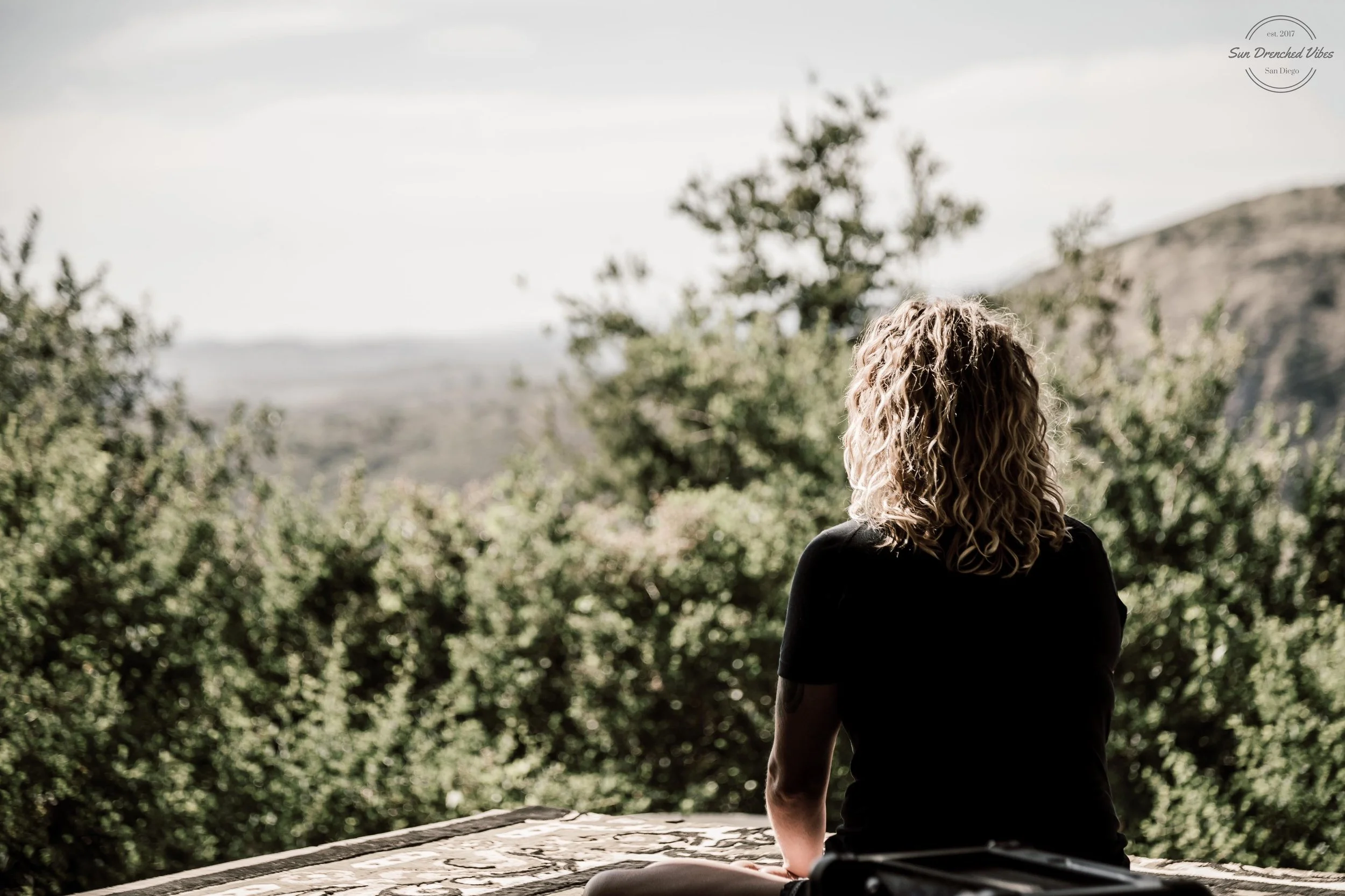 A person with curly blonde hair sitting on a stone ledge, overlooking a scenic landscape with trees and hills under a cloudy sky.