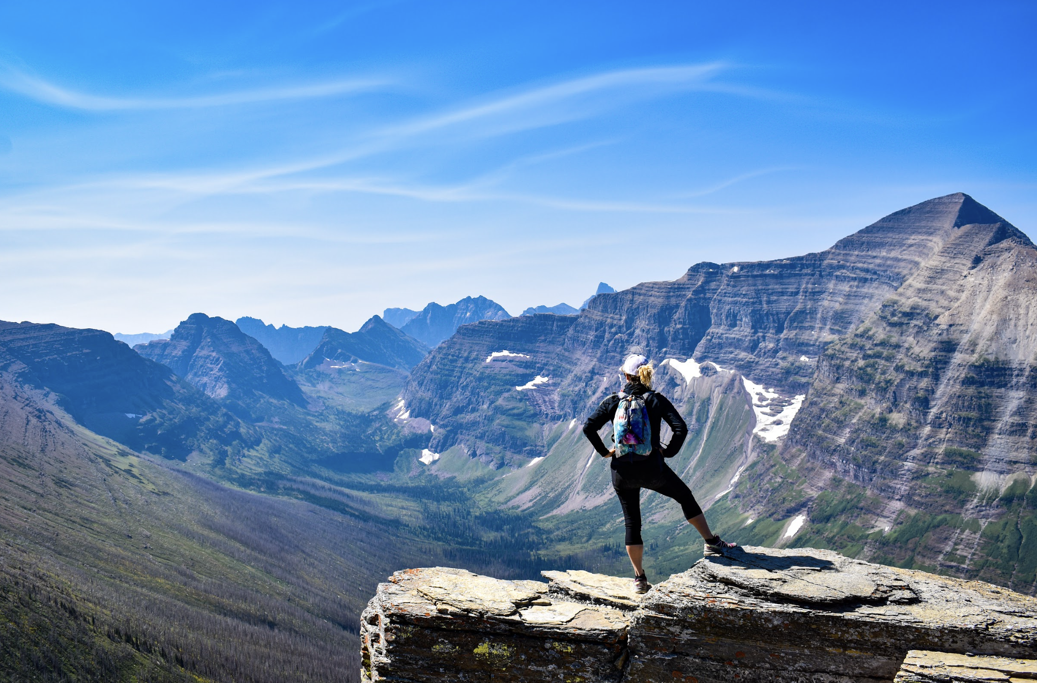Hiker standing on a rocky ledge overlooking a mountain range with snow patches under a blue sky.