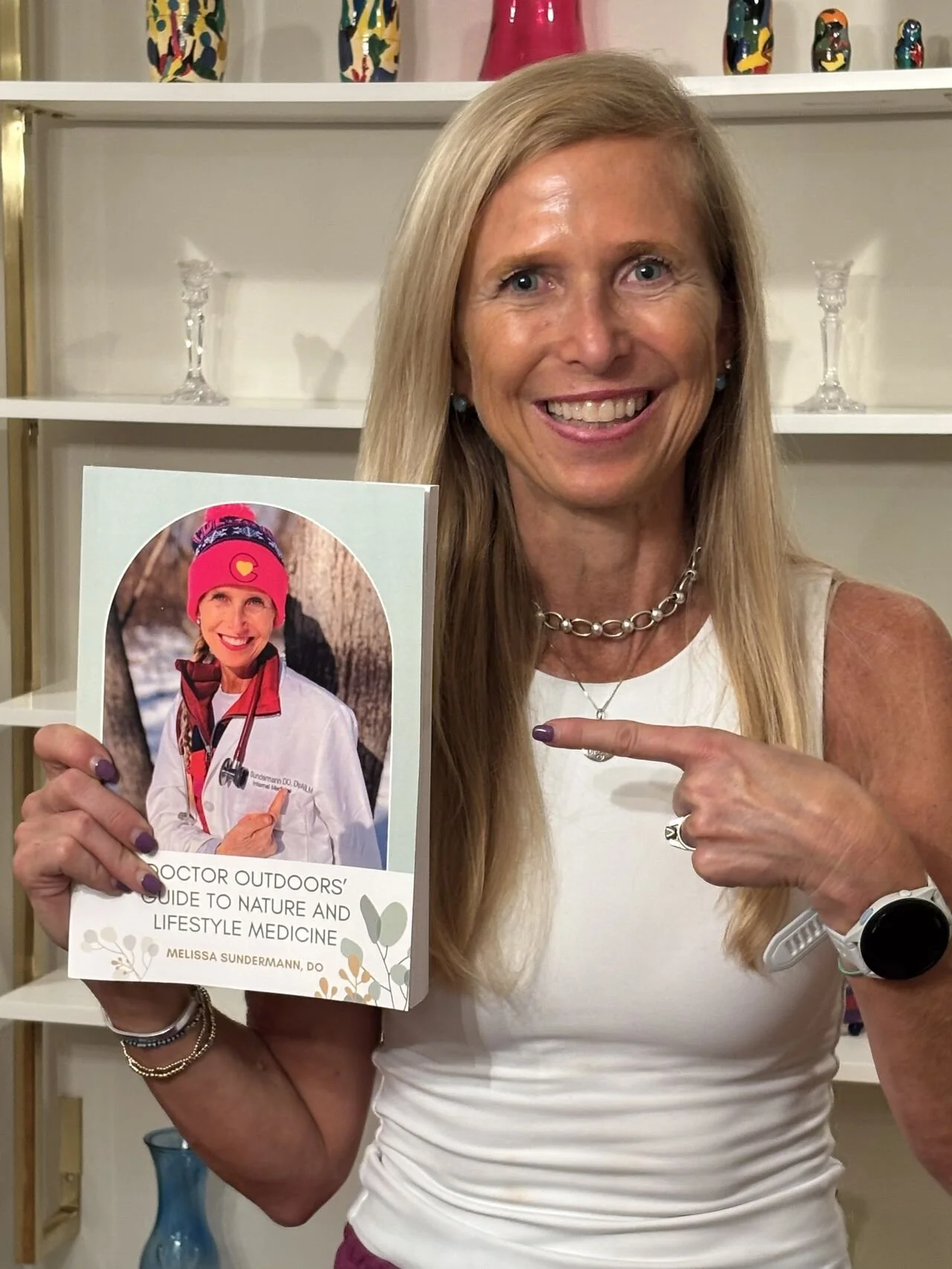 A smiling woman with long blonde hair holding a book titled 'Doctor Outdoors' Guide to Nature and Lifestyle Medicine' by Melissa Sundermann. She is pointing at the book with her left hand, wearing a white sleeveless top, a silver necklace, a watch, and bracelets, with a background of shelves with decorative items.