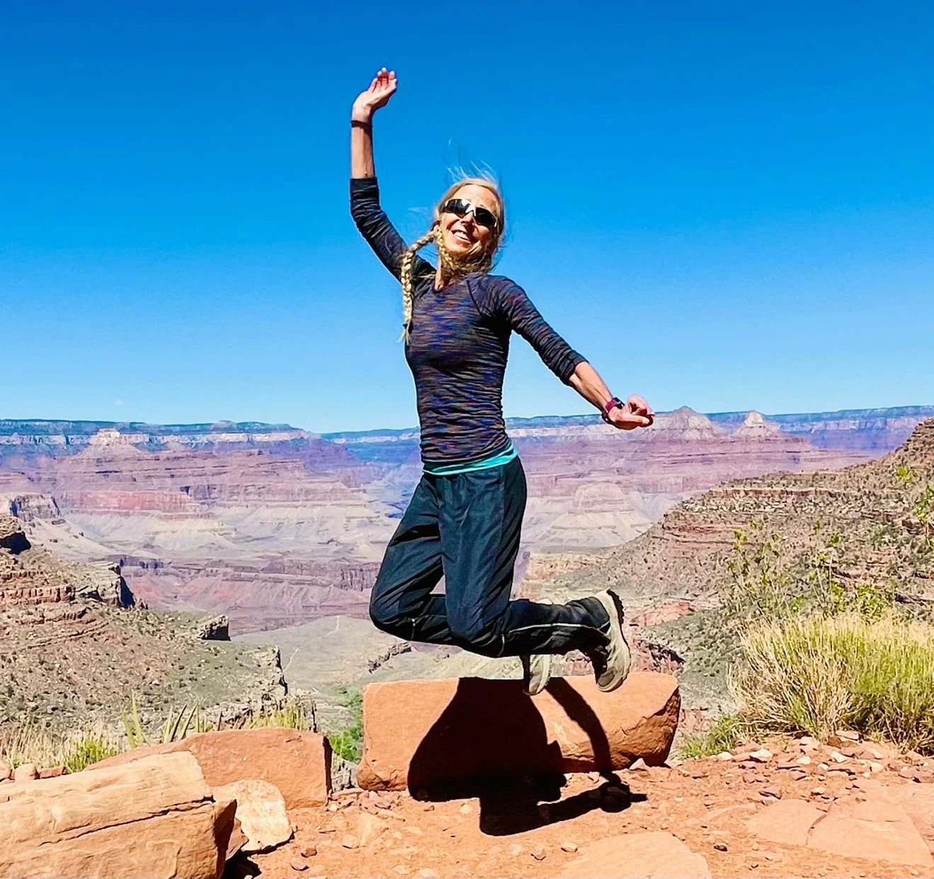 A woman jumping in the air with the Grand Canyon scenery in the background, wearing sunglasses, a long-sleeve athletic shirt, and athletic pants.