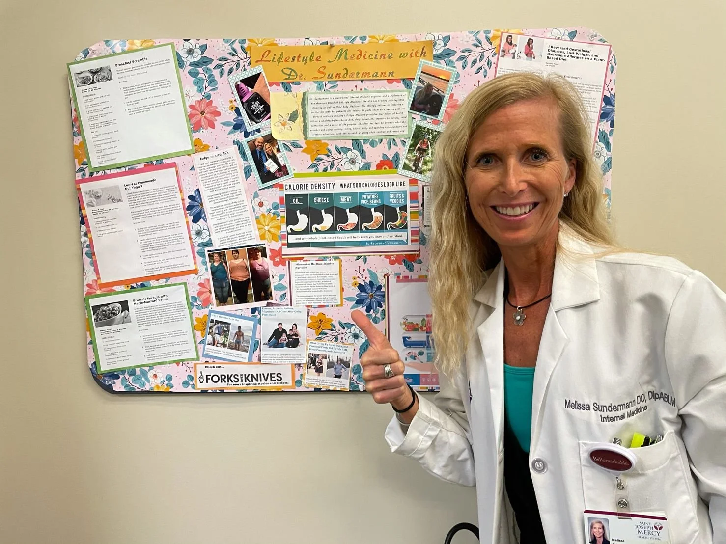 A woman in a white coat pointing at a colorful health and wellness poster on a bulletin board. The woman has blonde hair, is smiling, and wears a name tag that reads Melissa Sundermann, DO, Internal Medicine.