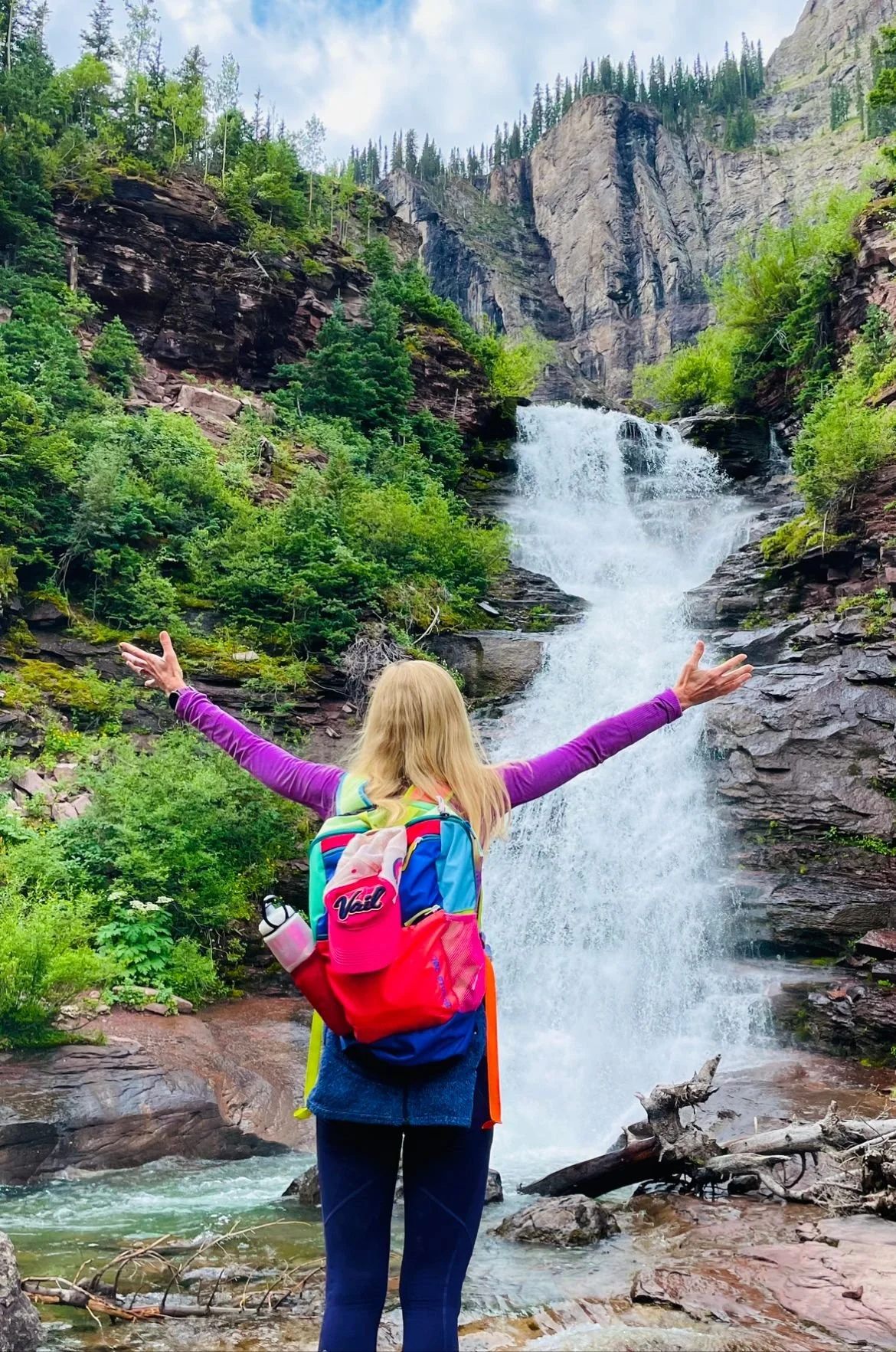 A woman with a backpack standing with arms outstretched in front of a waterfall in a green forested mountainous area.