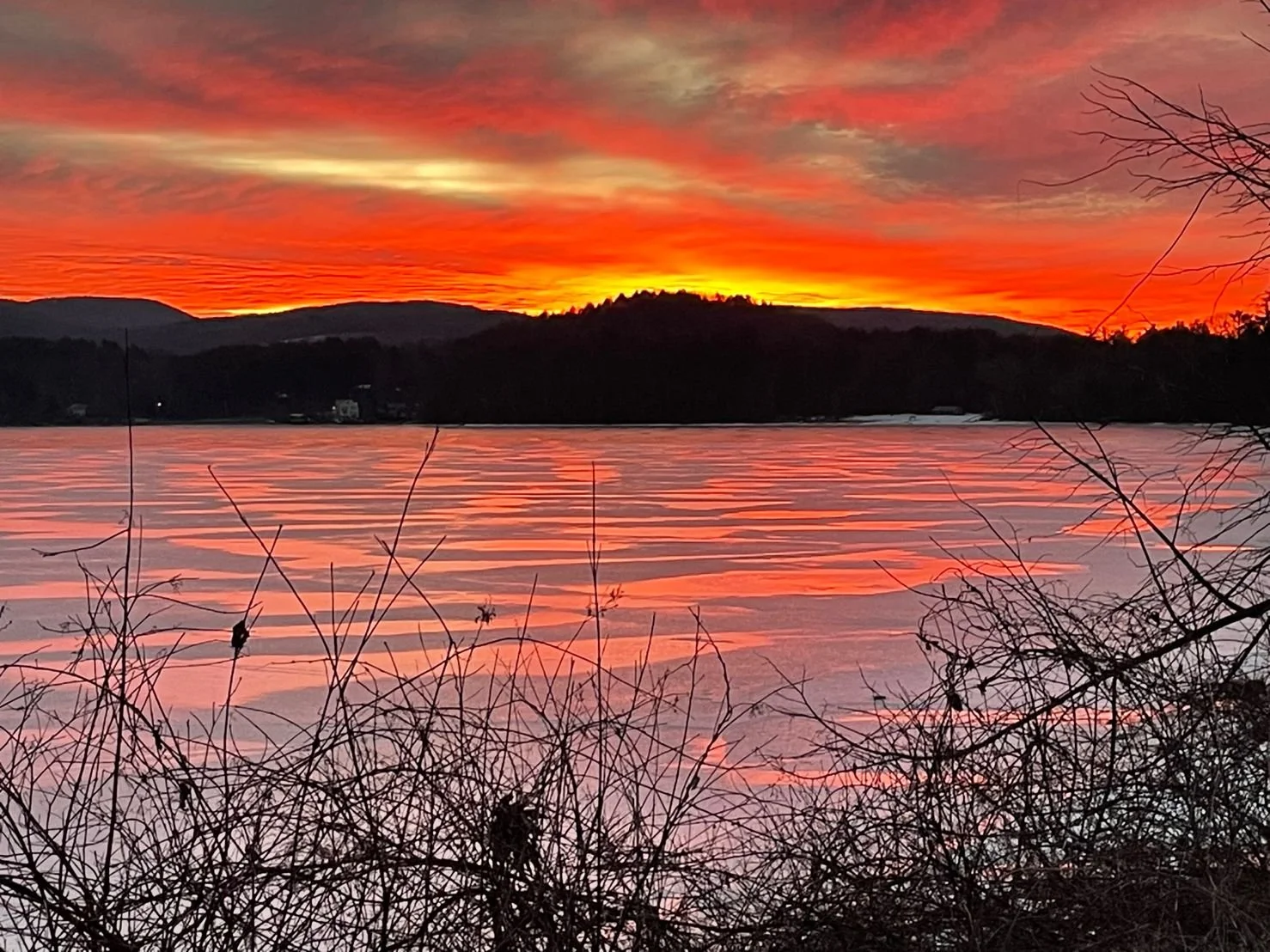 Sunset over a frozen lake with orange and pink clouds, silhouetted hills in the background, and leafless bushes in the foreground.