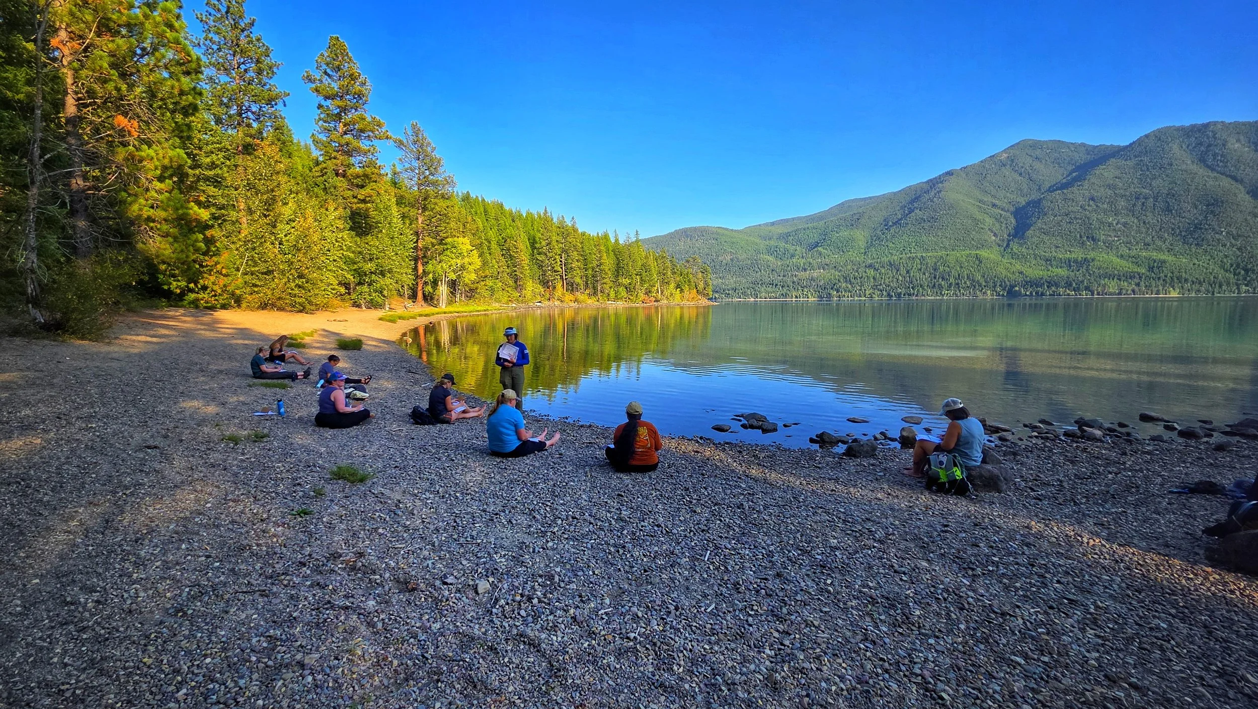 A group of people sitting on a pebbled beach by a lake surrounded by green trees and mountains, with a person standing by the water holding papers.