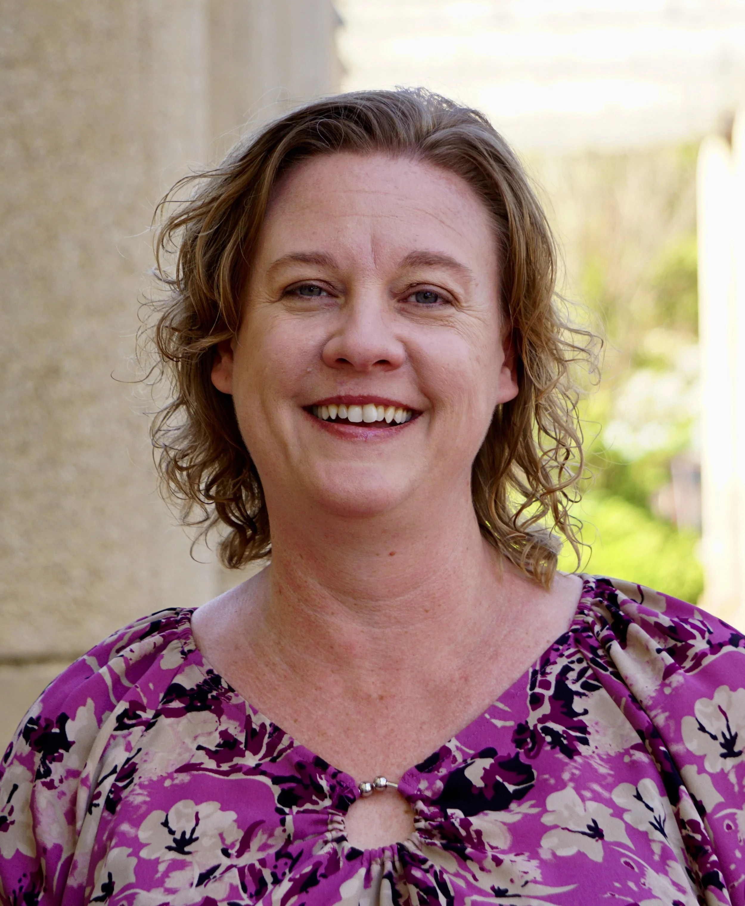 A woman with shoulder-length curly hair, smiling, wearing a purple patterned blouse with a round neckline and a small silver bead detail.