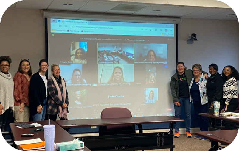 Group of ten women standing in a classroom with a large projection screen showing a Zoom video call in the background.