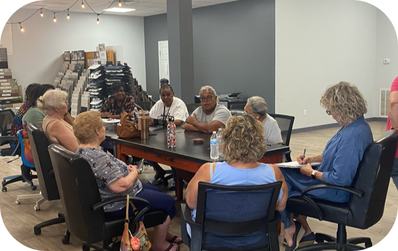 Group of women seated around a large conference table in a modern workspace, engaged in discussion. Some have notebooks, and there are water bottles on the table, with stacks of flooring samples in the background.