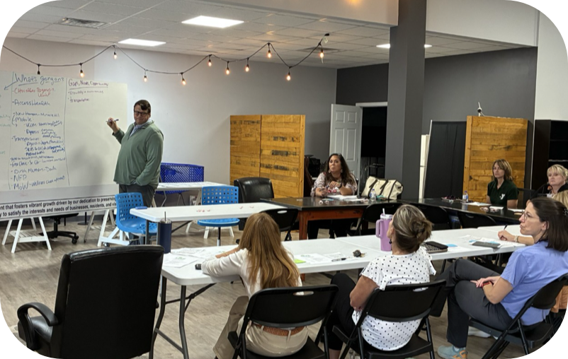 A man standing at a whiteboard with writing on it, giving a presentation to a group of women seated at tables in a classroom or conference room setting.