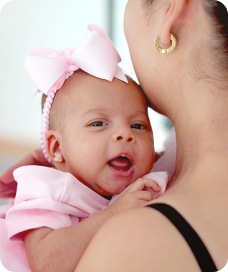 A baby girl wearing a pink dress and headband with a large pink bow, being held close to her mother, who has gold earrings and is wearing a black strap top.