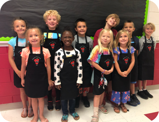 Ten smiling children, wearing black aprons with a red apple logo, standing together inside a classroom with red and green walls.