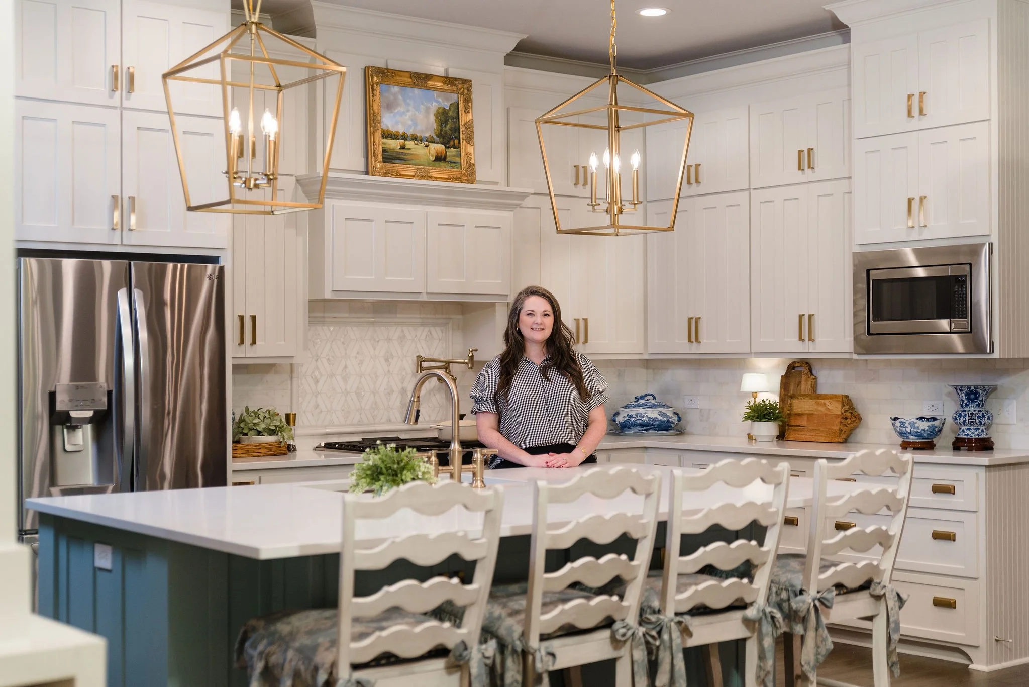 A woman standing in a modern, well-lit kitchen with white cabinetry, gold hardware, and a white island. The kitchen features a stainless steel refrigerator, microwave, and a gold pendant light above the island. There are decorative blue and white dishes and plants on the counter.