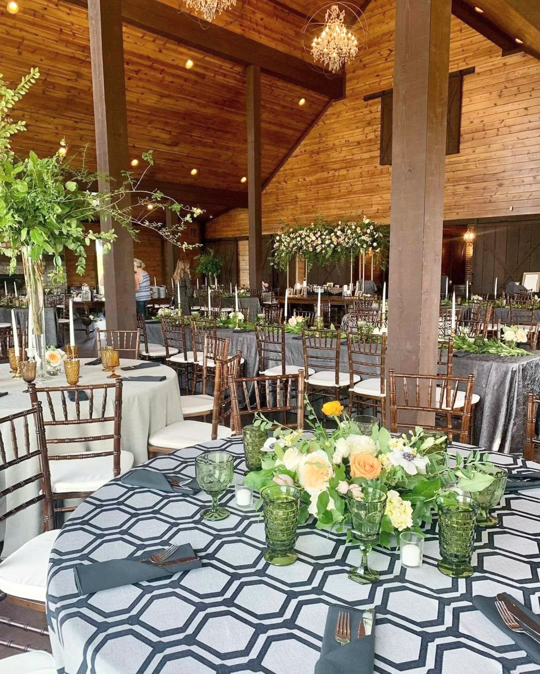 Elegant event space with round tables decorated with floral centerpieces, green glassware, and black napkins, under a wooden ceiling with chandeliers.