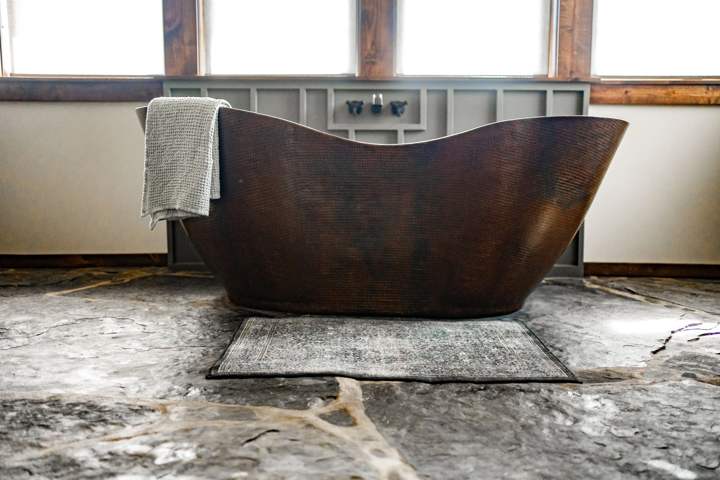 A rustic bathroom featuring a large, dark wooden bathtub with a textured surface, placed on a stone floor with a small patterned rug underneath. A gray towel hangs over the edge of the tub, and the background shows a wall with three small black decorative objects and three large windows.