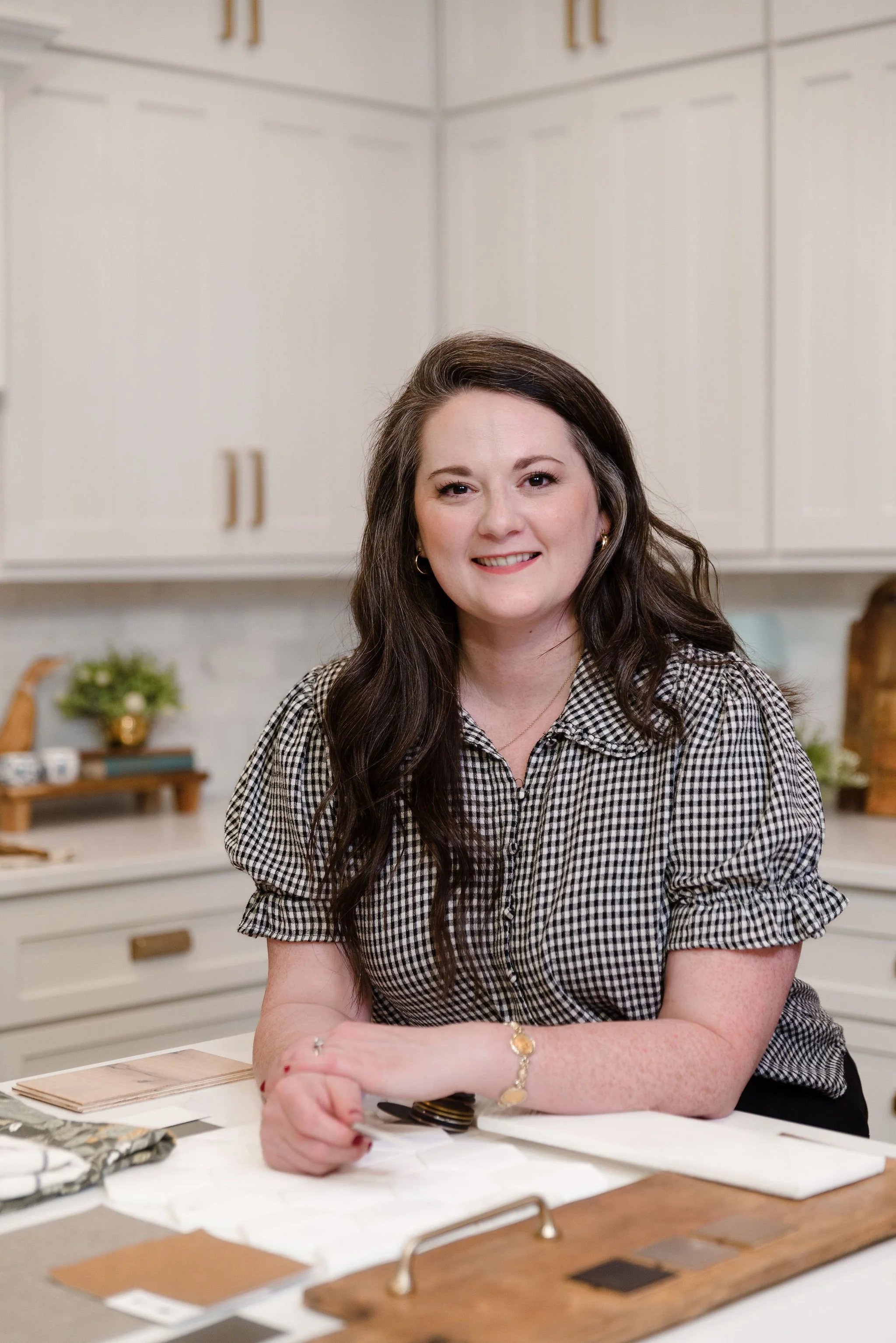 A woman with long dark hair and fair skin smiles at the camera in a modern kitchen. She is wearing a black and white checkered shirt and gold jewelry, with various fabric samples and color swatches on the table in front of her.