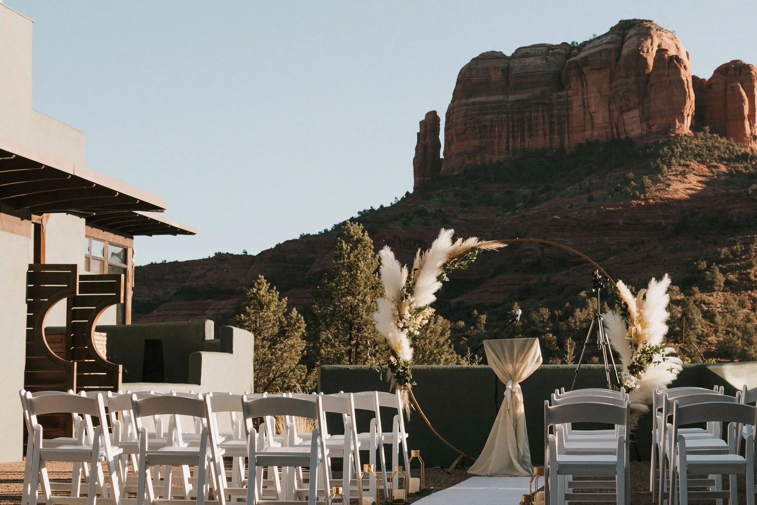 Outdoor wedding setup with white chairs, floral arrangements, and a circular arch, overlooking a mountain with red rock formations.