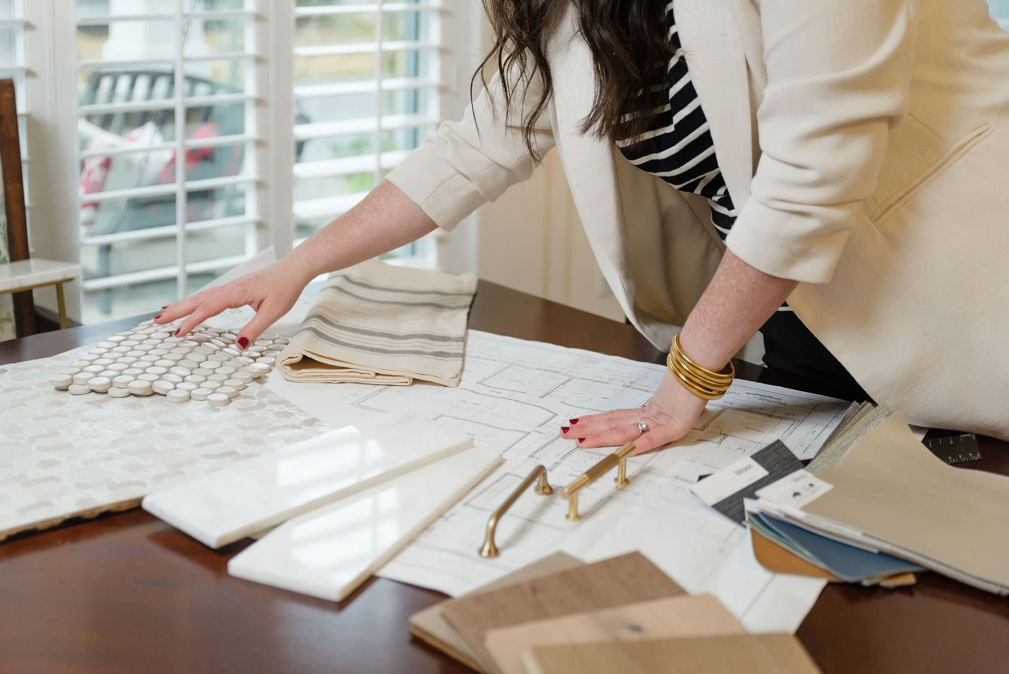 A person working on interior design or architecture plans at a desk, surrounded by fabric samples, tile samples, and color swatches, in a well-lit room with large windows.