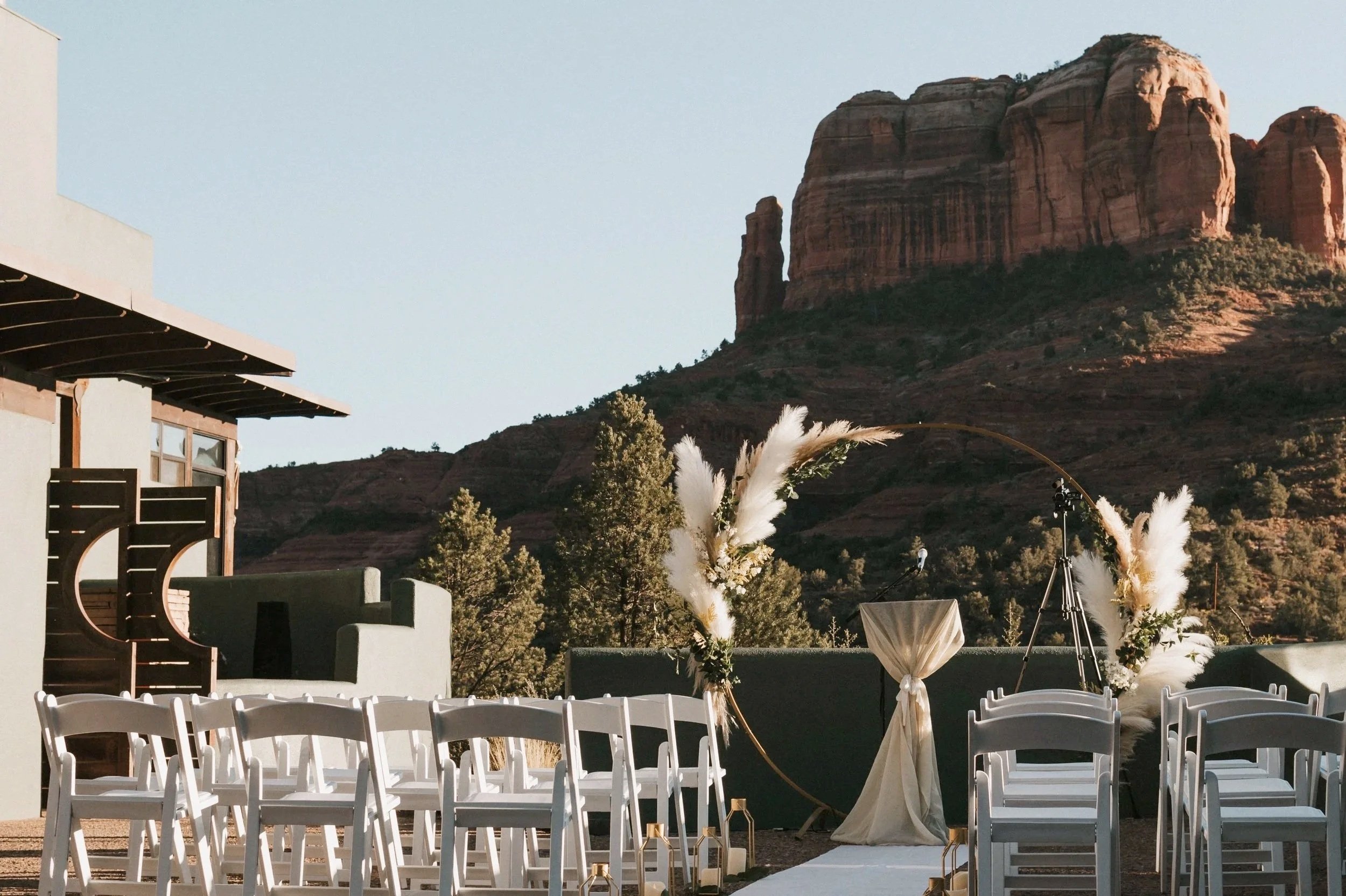 Outdoor wedding setup with white chairs, a floral arch with pampas grass, and a mountain landscape in the background.