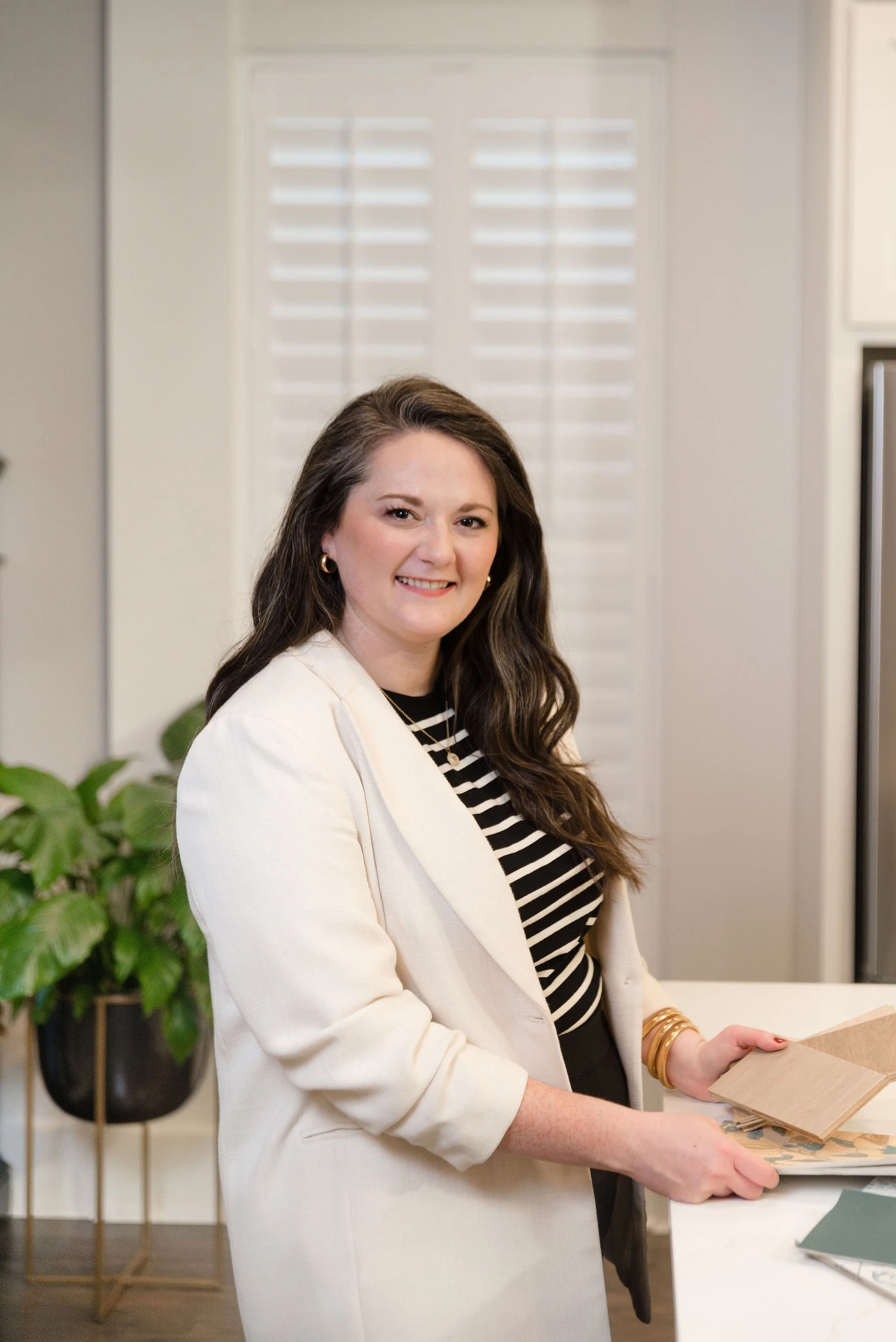 A woman with long brown hair and gold hoop earrings smiling in a kitchen, wearing a white blazer over a black and white striped shirt, holding different samples or swatches.