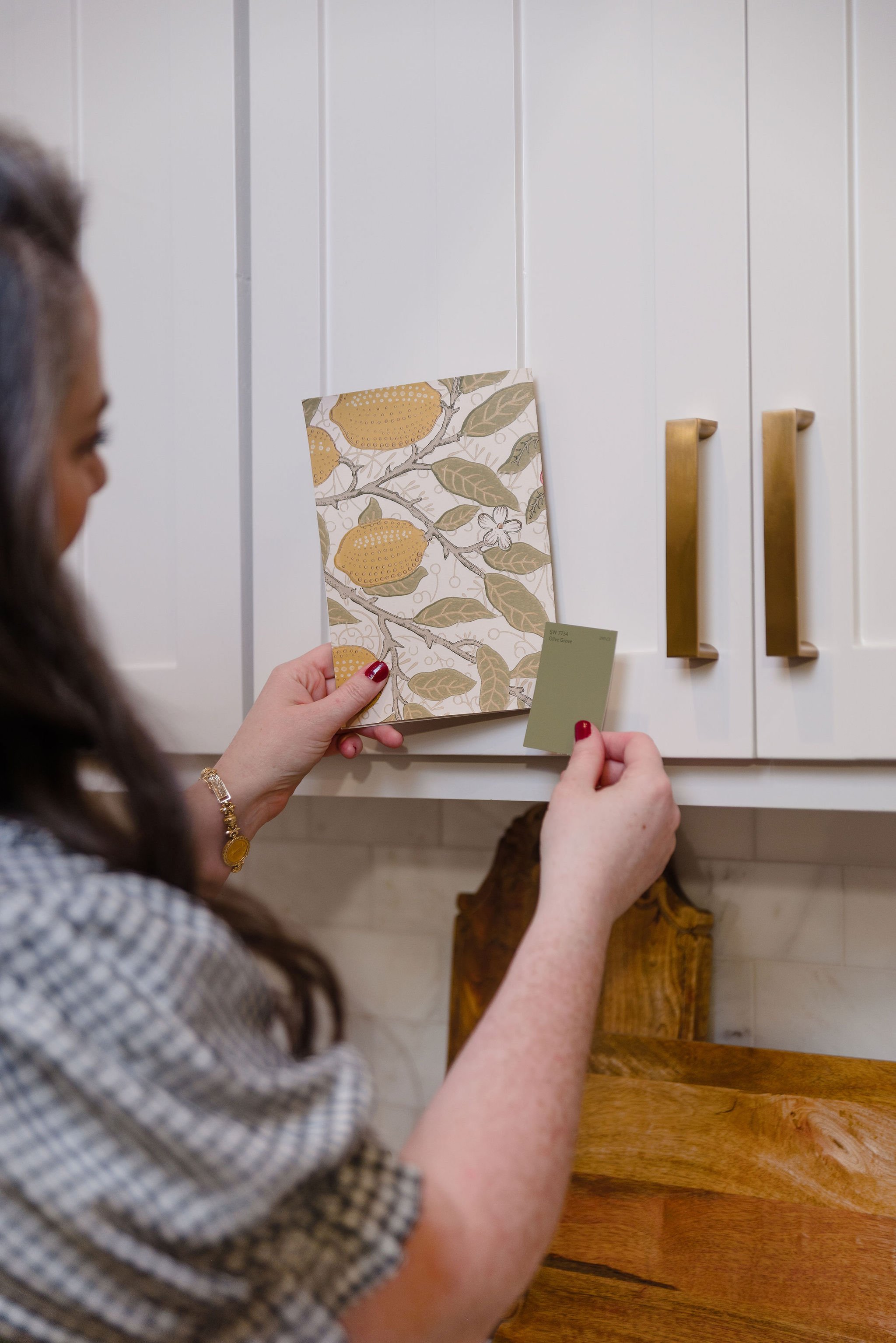 A woman holding two sample paint color cards in front of white kitchen cabinets with gold handles, with a wooden cutting board below.