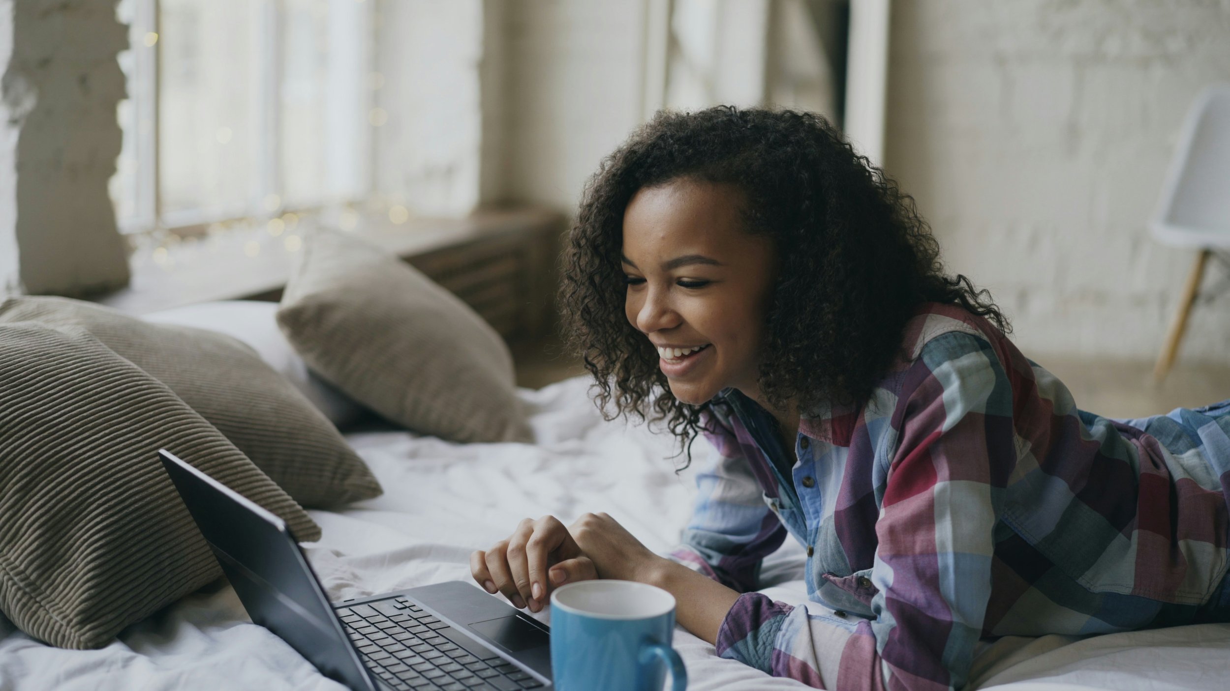 A woman lying on her stomach on a bed, looking at a laptop and smiling in a bright, cozy room with large windows.