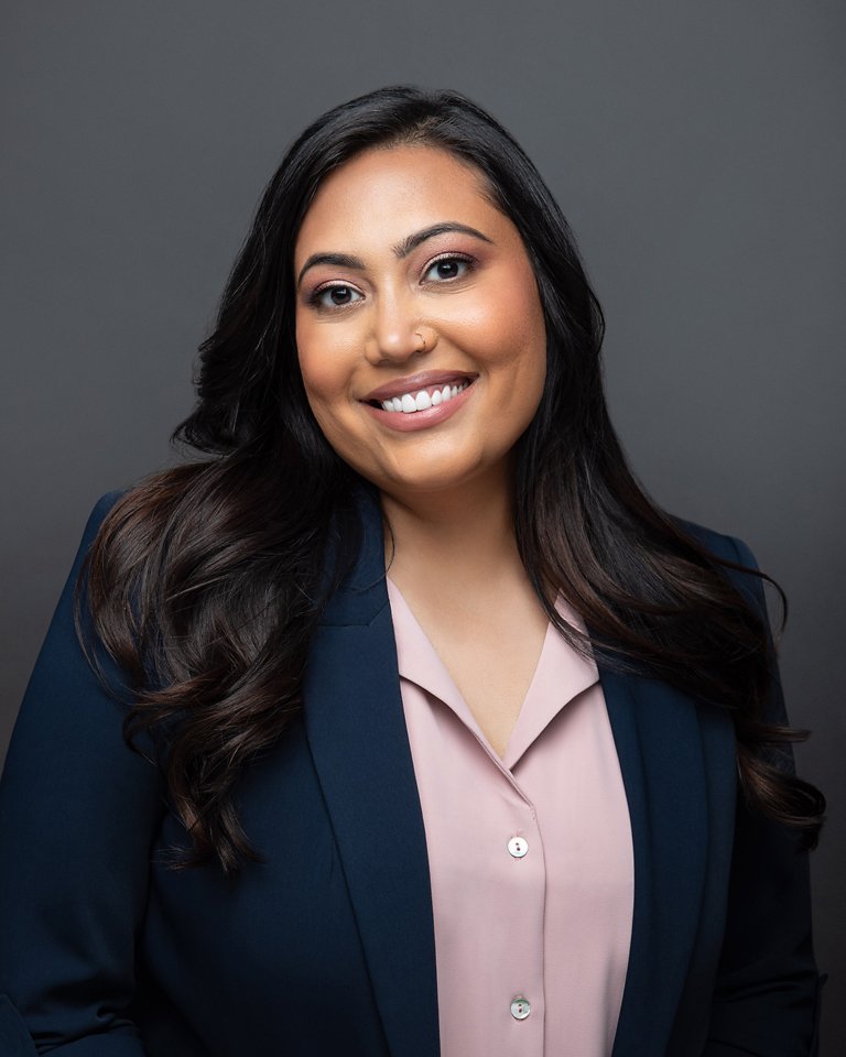 A woman with long dark hair, wearing a navy blazer and light pink blouse, smiling against a gray background.