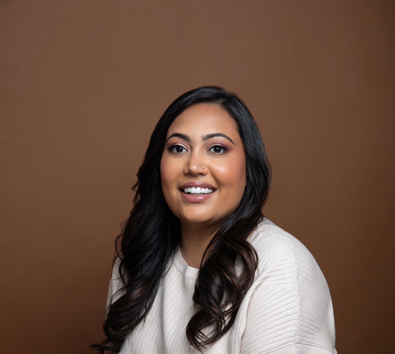 A woman with long dark hair and a light-colored sweater, smiling at the camera, against a brown background.