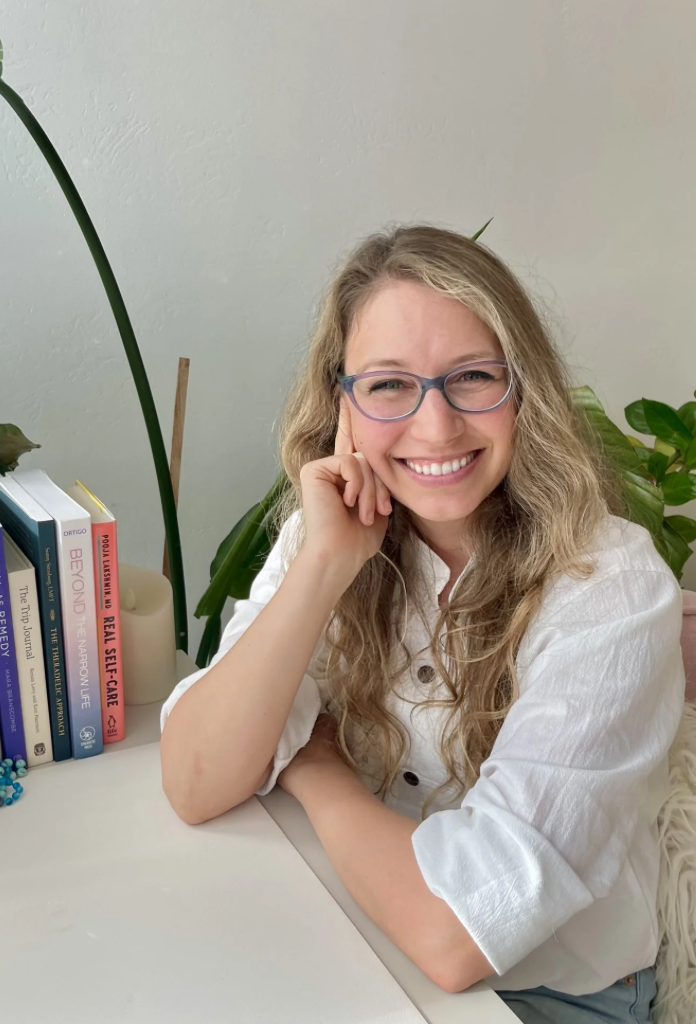 A woman with curly blonde hair, glasses, and a white shirt smiling at the camera, sitting at a desk with books and green plants in the background.