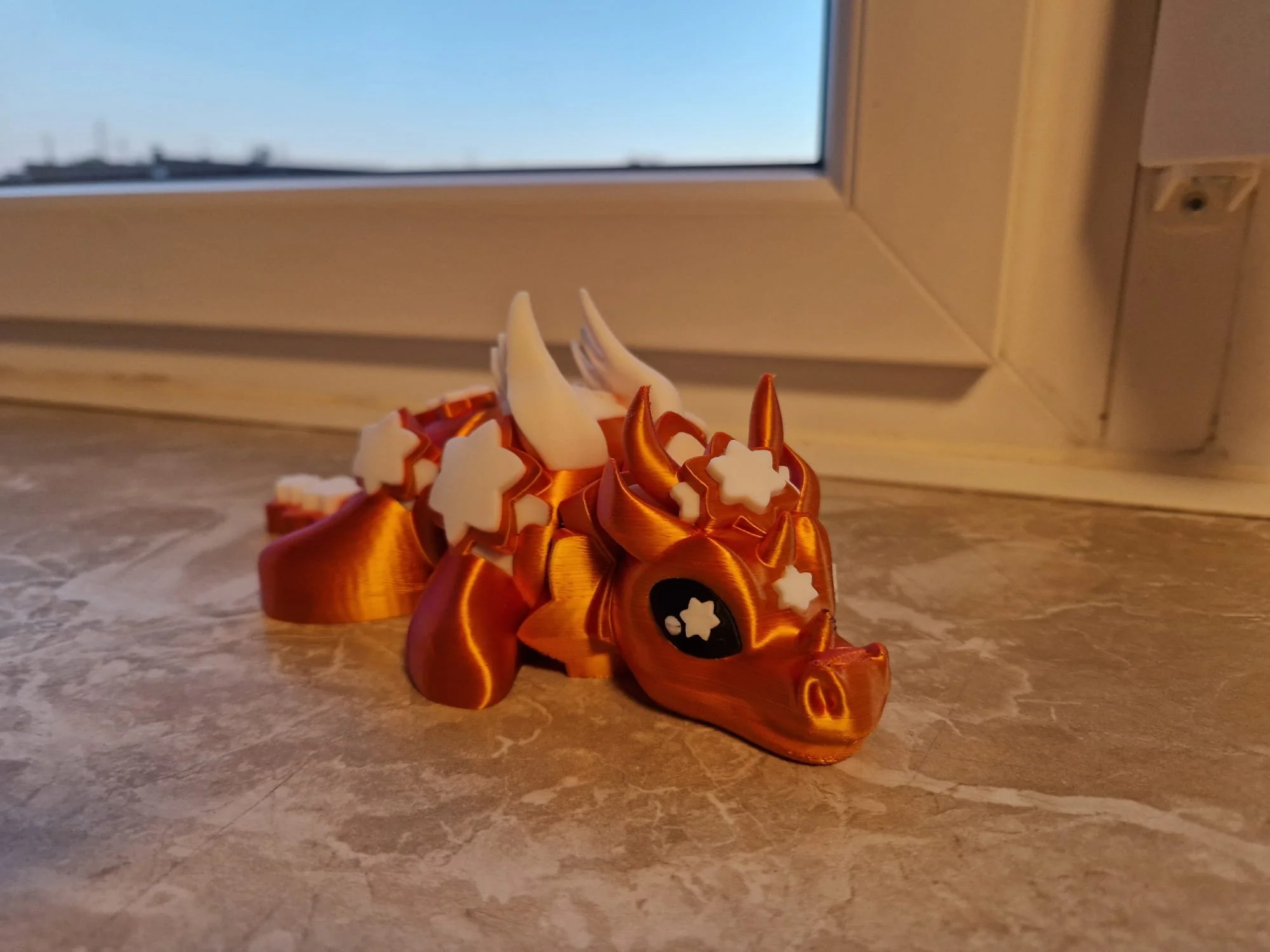 Ein 3D-gedruckter organgener Flexi-Drache mit Hörnern und Sternen. Der Druck ist mehrfarbig.