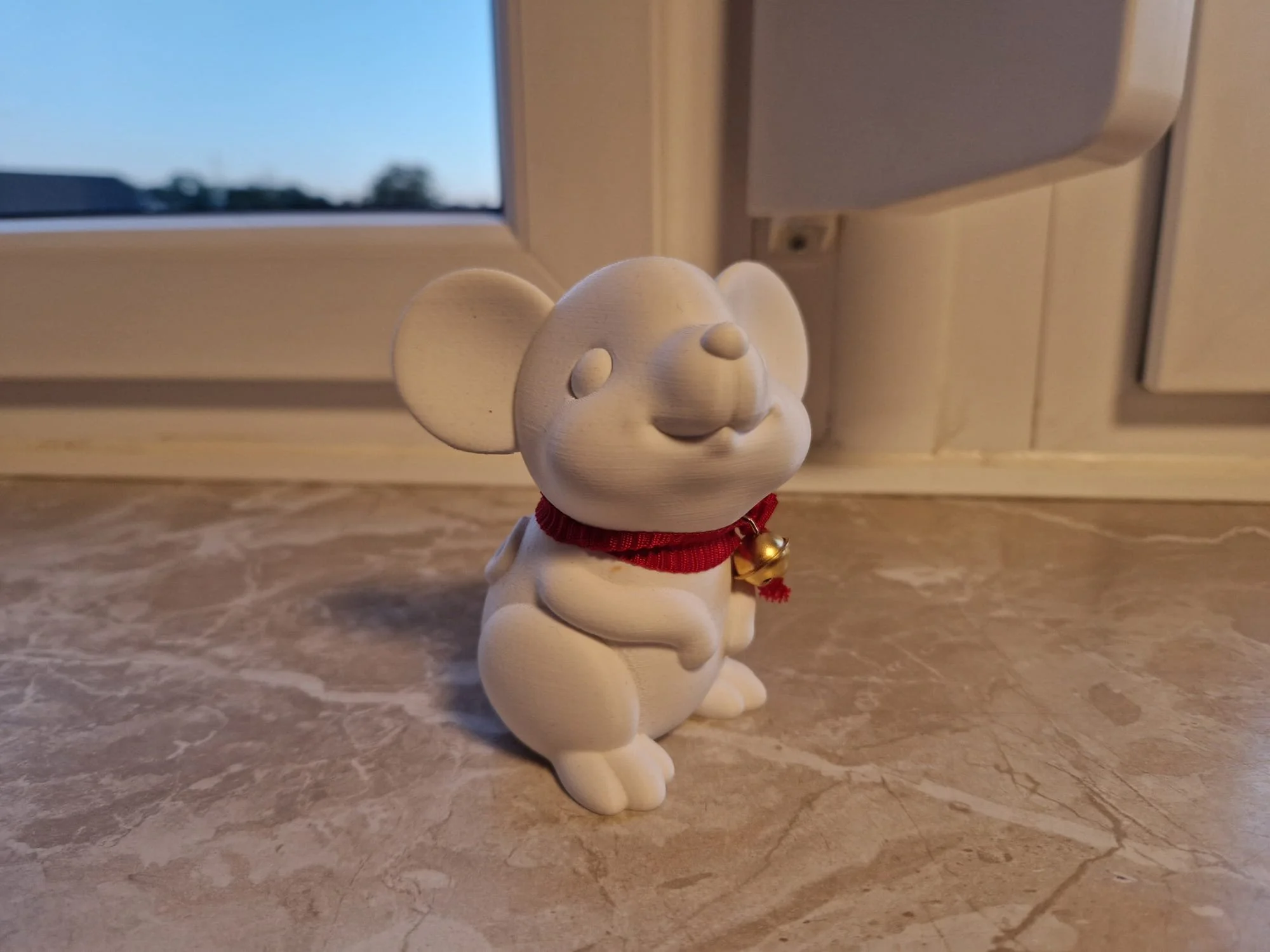 Eine 3D-gedruckte weiße Maus mit großen Ohren und einem roten Halsband mit Glöckchen. 