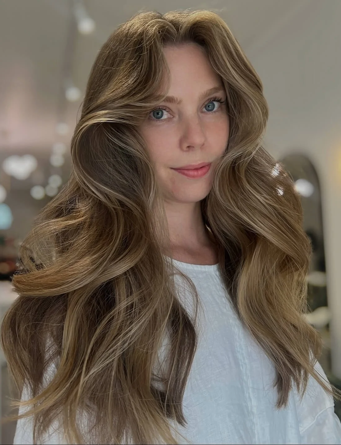 A woman with long, wavy, light brown hair and blue eyes, looking directly at the camera with a slight smile, in an indoor setting.