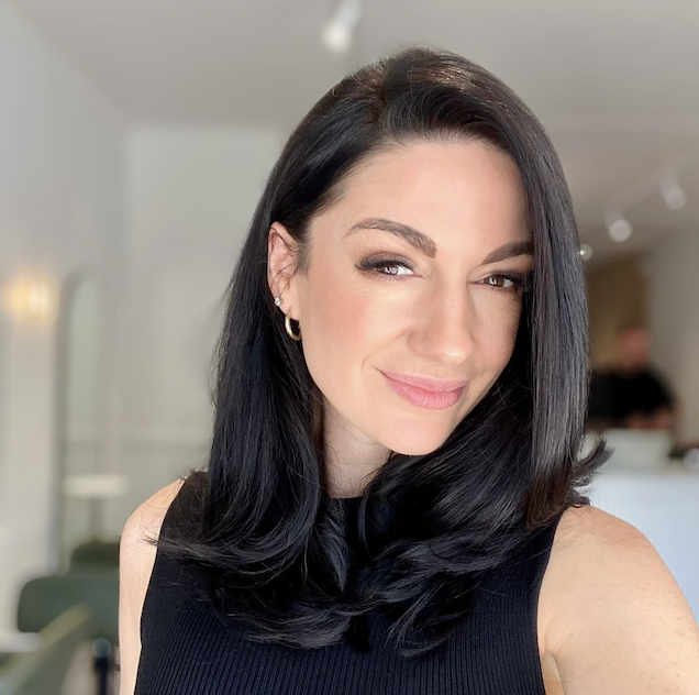 Close-up of a woman with long, wavy, blonde hair and blue eyes, wearing a white top, in an indoor setting.